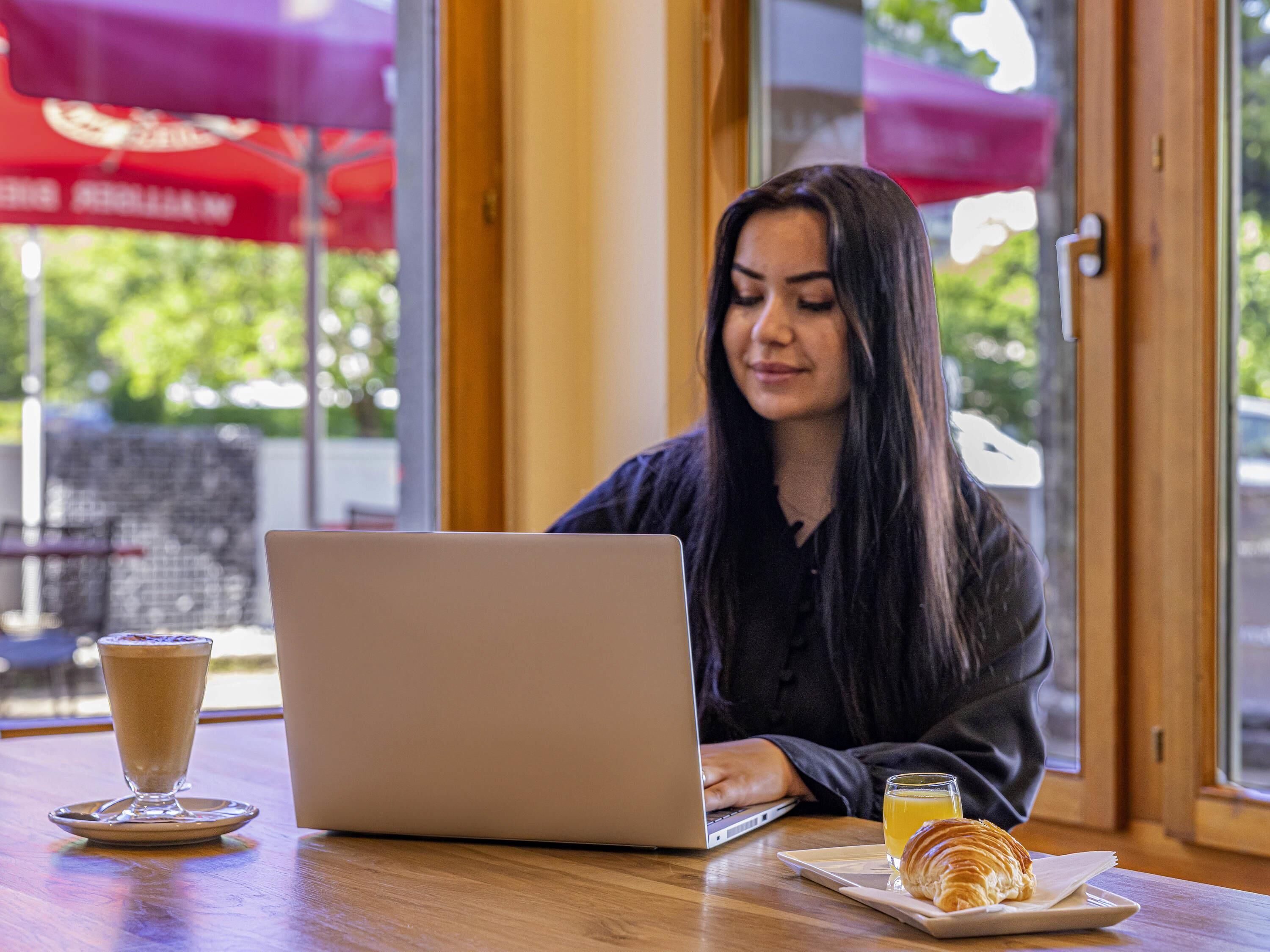 Una persona usando una computadora portátil en una mesa, con una bebida y un pastel.
