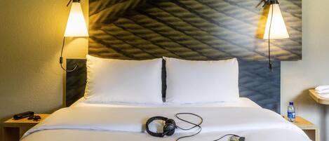 A neatly made bed with white linens, a pair of headphones, a book, and a pair of glasses on it. There are two bedside tables with lamps, one on each side of the bed.