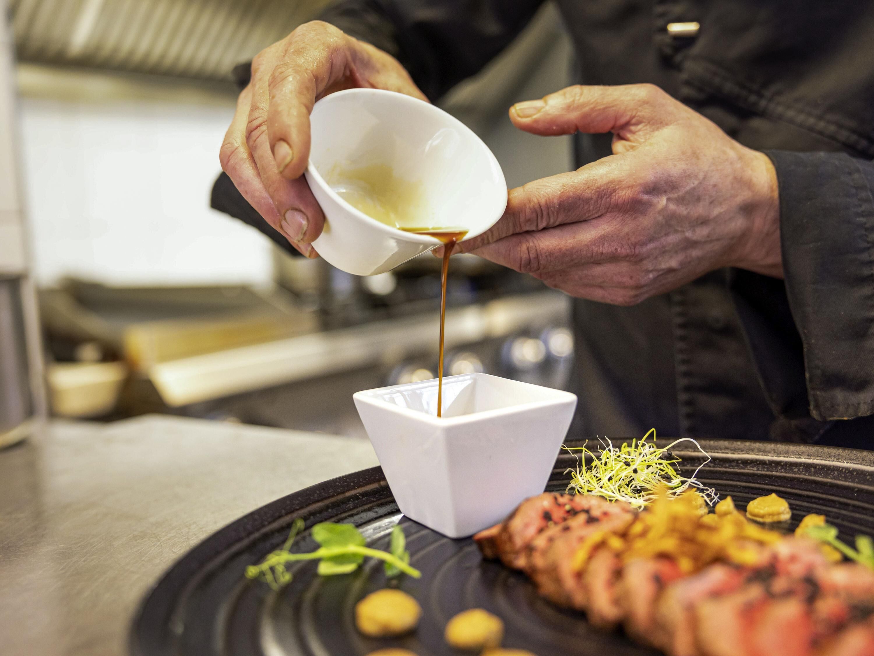 Une personne verse de la sauce dans un bol blanc posé sur une assiette contenant de la nourriture.