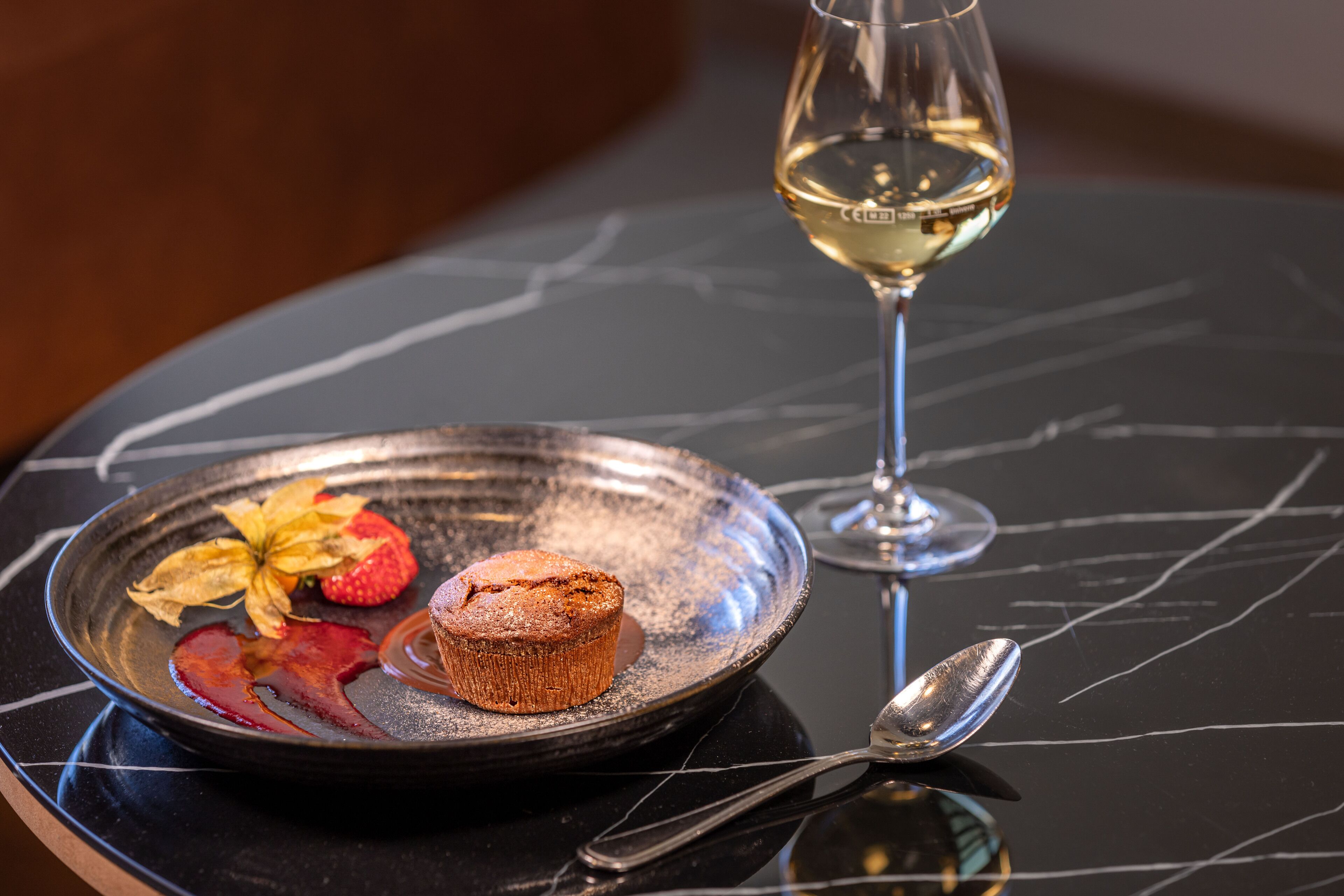 A dessert plate with a muffin and a strawberry on a black marble table, accompanied by a wine glass and a spoon.