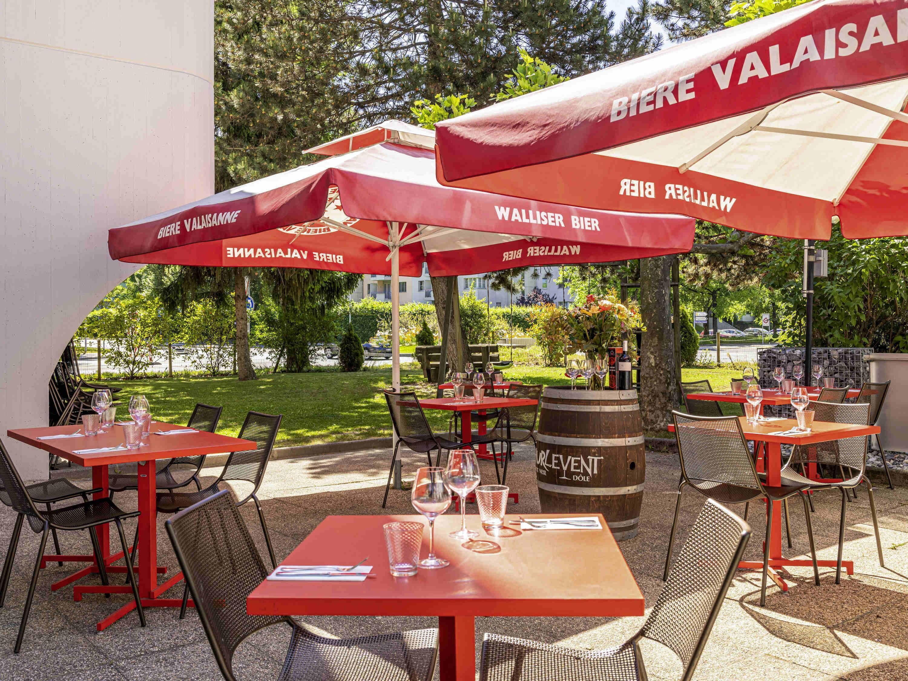 Outdoor tables and chairs with red umbrellas, a wooden barrel, and a view of greenery and buildings.