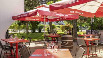 Outdoor tables and chairs with red umbrellas, a wooden barrel, and a view of greenery and buildings.