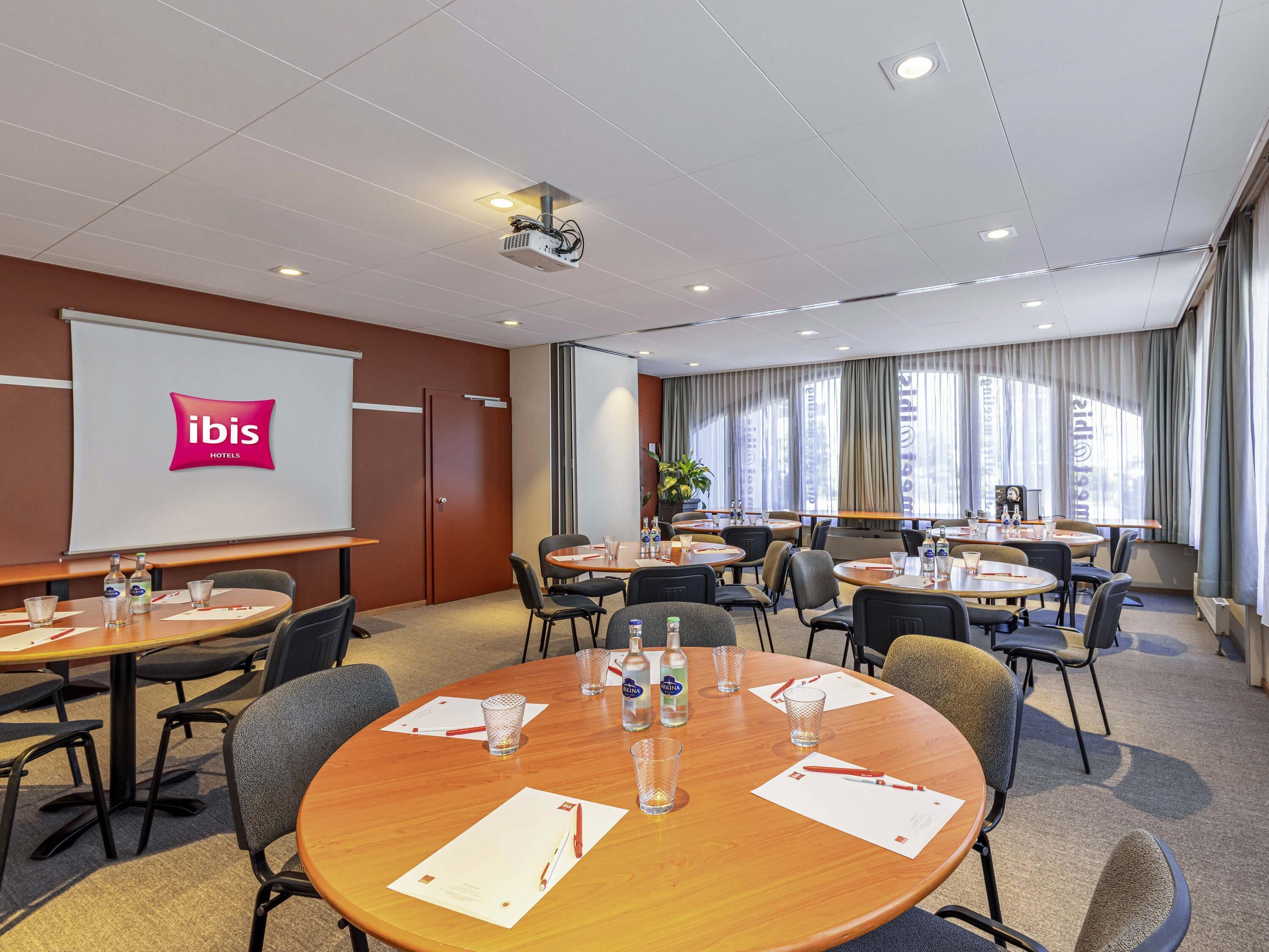 A conference room with a projector, a screen displaying the ibis logo, multiple tables with chairs, and a large window with curtains.