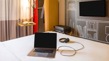 Ein Hotelzimmer mit einem Bett, einem Laptop, Kopfhörern, einem Koffer, einem Fernseher und einer Wand mit Kreidezeichnungen.