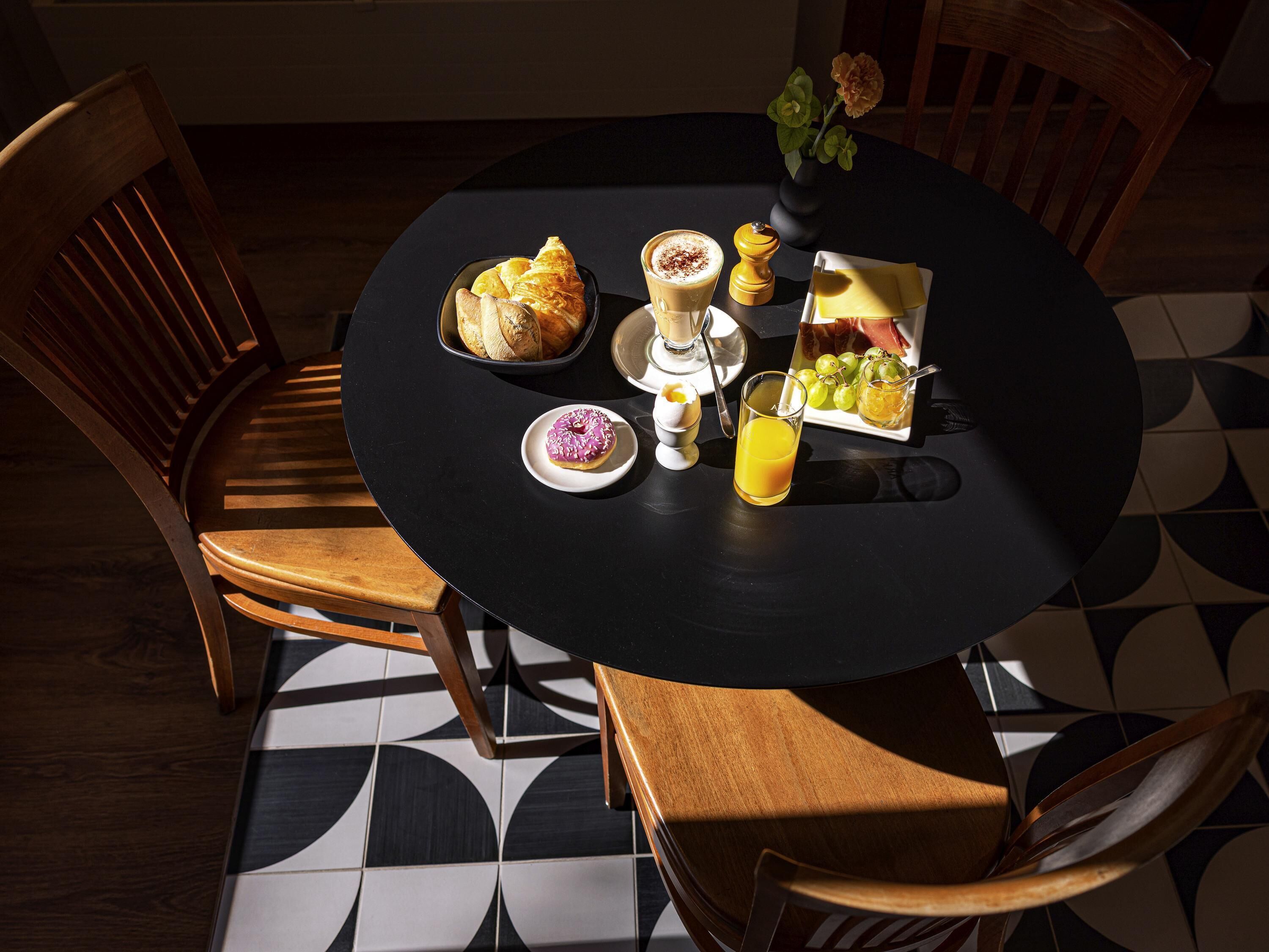 A round dining table with a black tablecloth, surrounded by wooden chairs. On the table, there is a plate with croissants, a plate with a pink frosted donut, a glass of orange juice, a small bowl with a white substance, and a small vase with flowers.