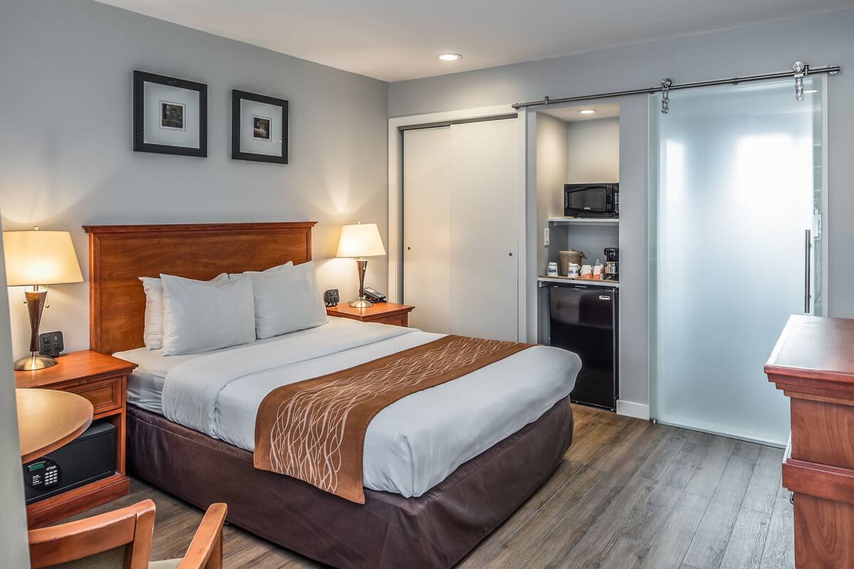 A hotel room with a bed, bedside lamps, a desk, a chair, a microwave, a refrigerator, and a sliding door closet.
