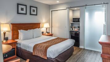 A hotel room with a bed, bedside lamps, a desk, a chair, a microwave, a refrigerator, and a sliding door closet.