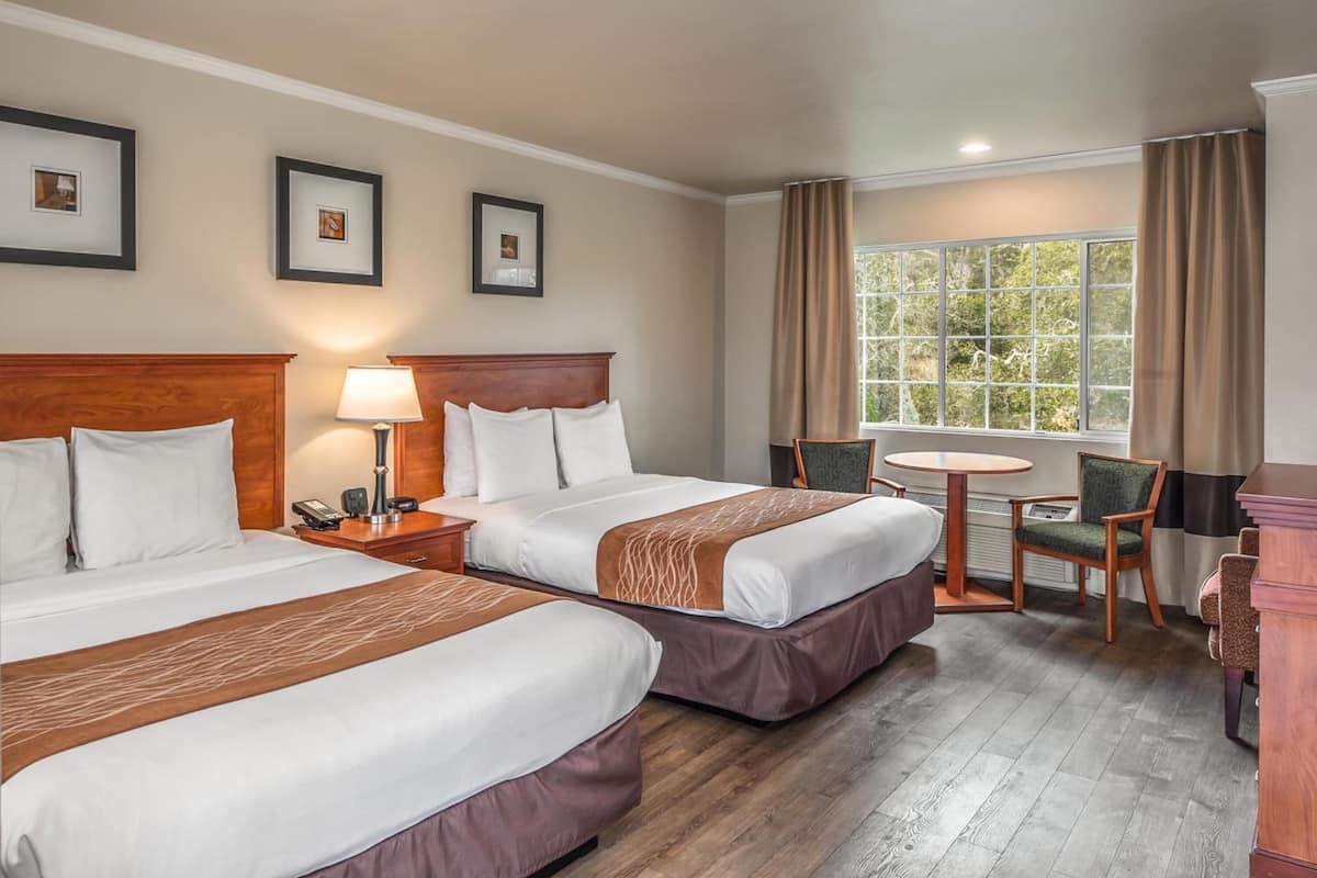 A hotel room with two beds, a desk, a chair, a small table, and a window with curtains.