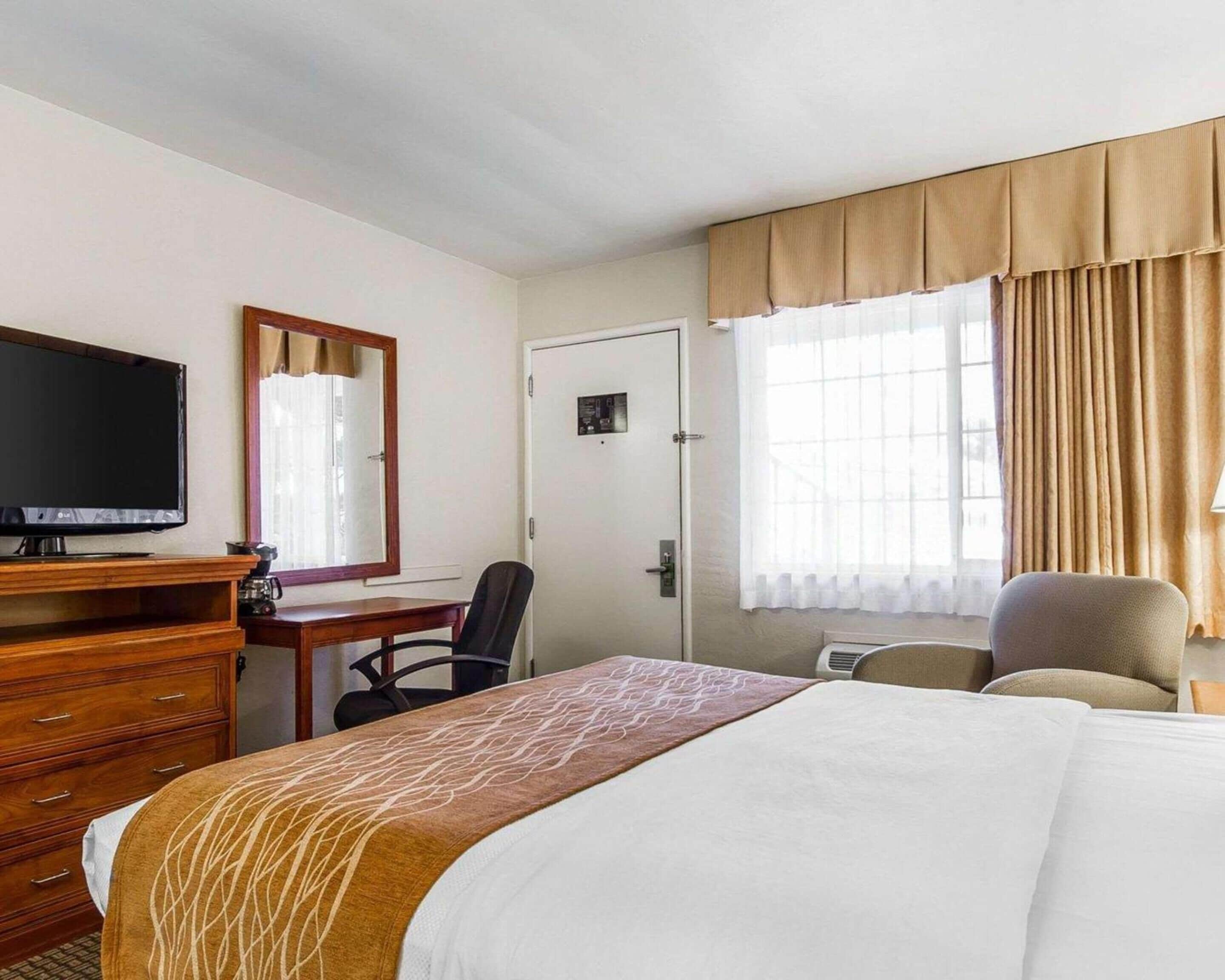 Una habitación de hotel con cama, escritorio con televisor, una silla y ventana con cortinas.