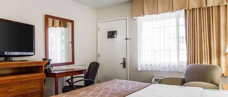 Una habitación de hotel con cama, escritorio con televisor, una silla y ventana con cortinas.