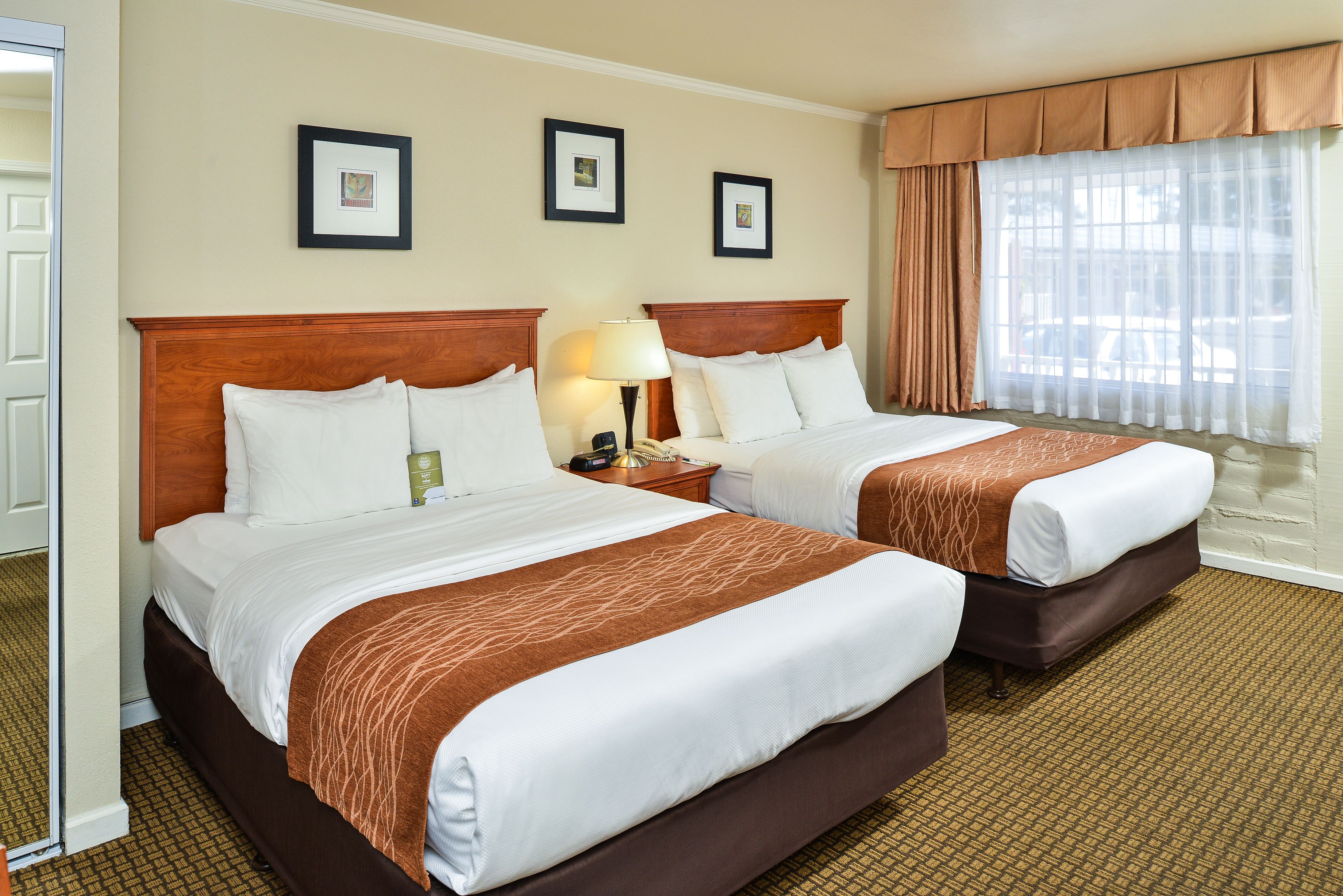 Quarto de hotel com duas camas, cabeceira de madeira, mesa de cabeceira com abajur e janela com cortinas leves.