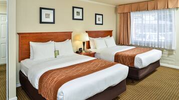 Quarto de hotel com duas camas, cabeceira de madeira, mesa de cabeceira com abajur e janela com cortinas leves.