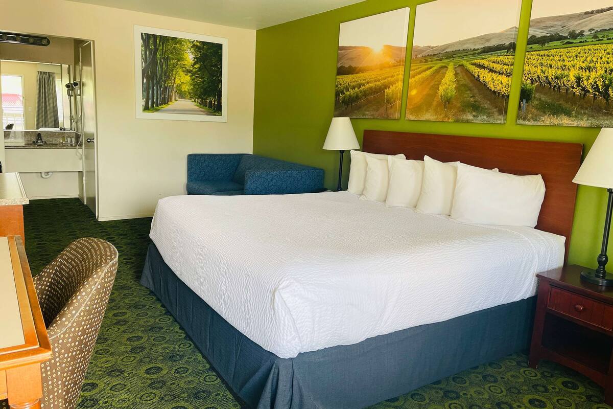 A hotel room with a large bed, a desk with a chair, a mirror, and two paintings on the wall.