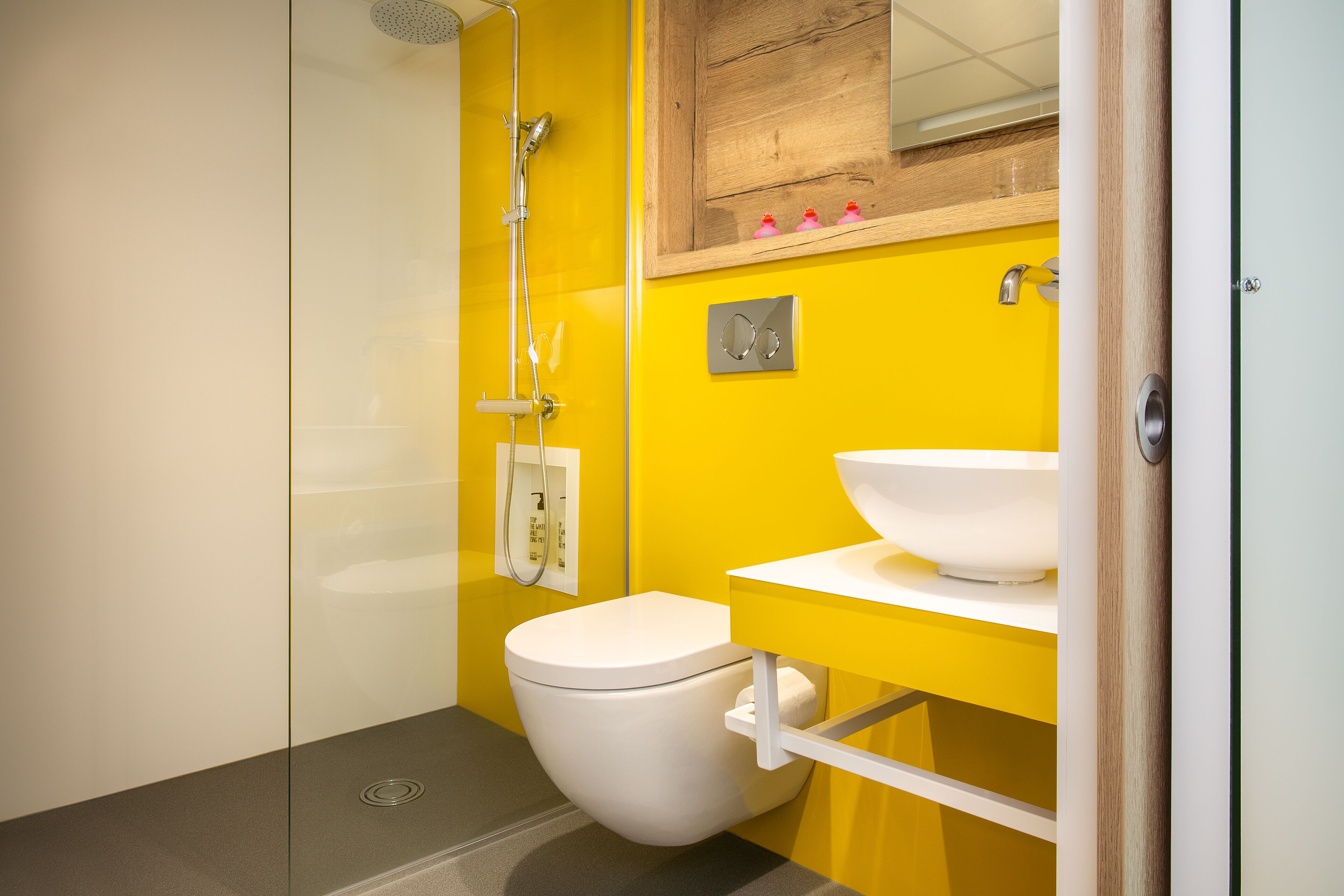 A modern bathroom with a yellow wall, a white toilet, a white sink, and a shower.