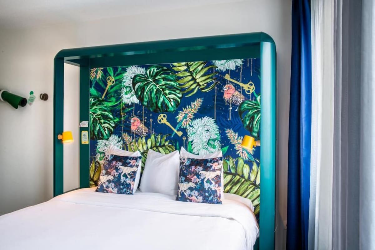 Un lit doté d’une tête de lit ornée d’un motif de feuilles tropicales, d’un couvre-lit blanc et de deux coussins décoratifs.
