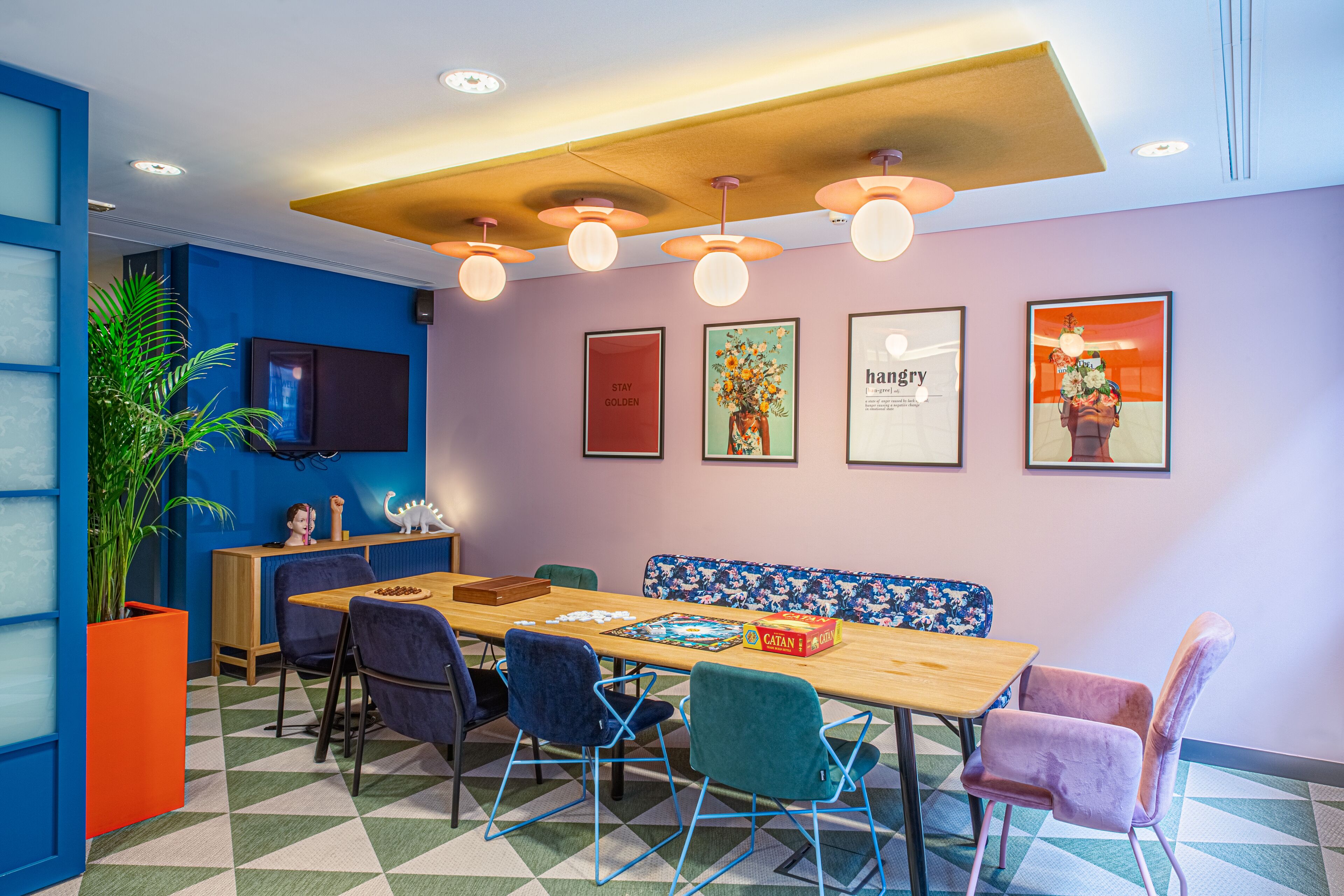 Un comedor moderno con una mesa de madera, sillas azules y moradas, y un banco. Hay cuadros en la pared y una planta en maceta.