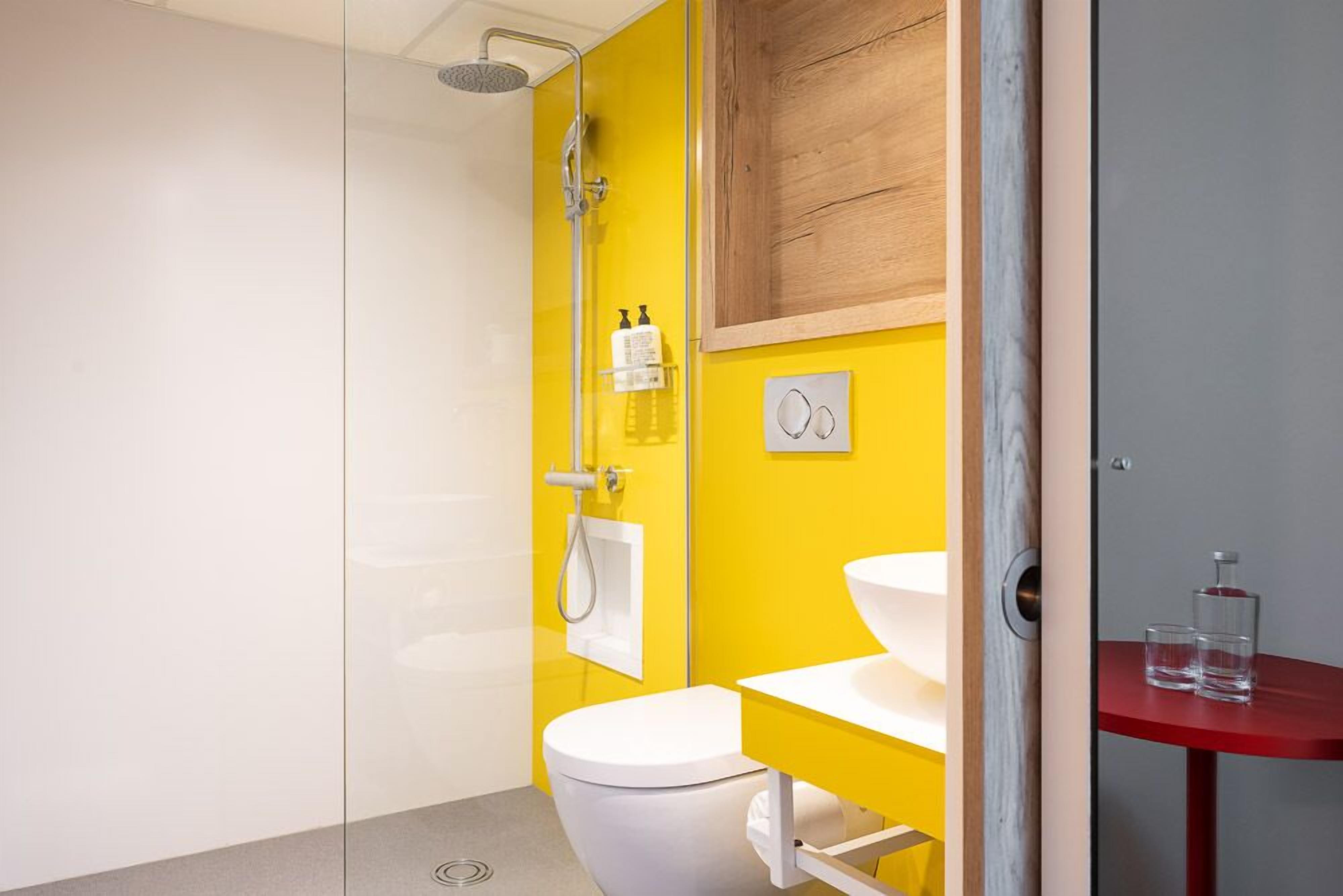 Une salle de bain avec un mur jaune, des toilettes blanches, une douche en verre et un lavabo rouge.