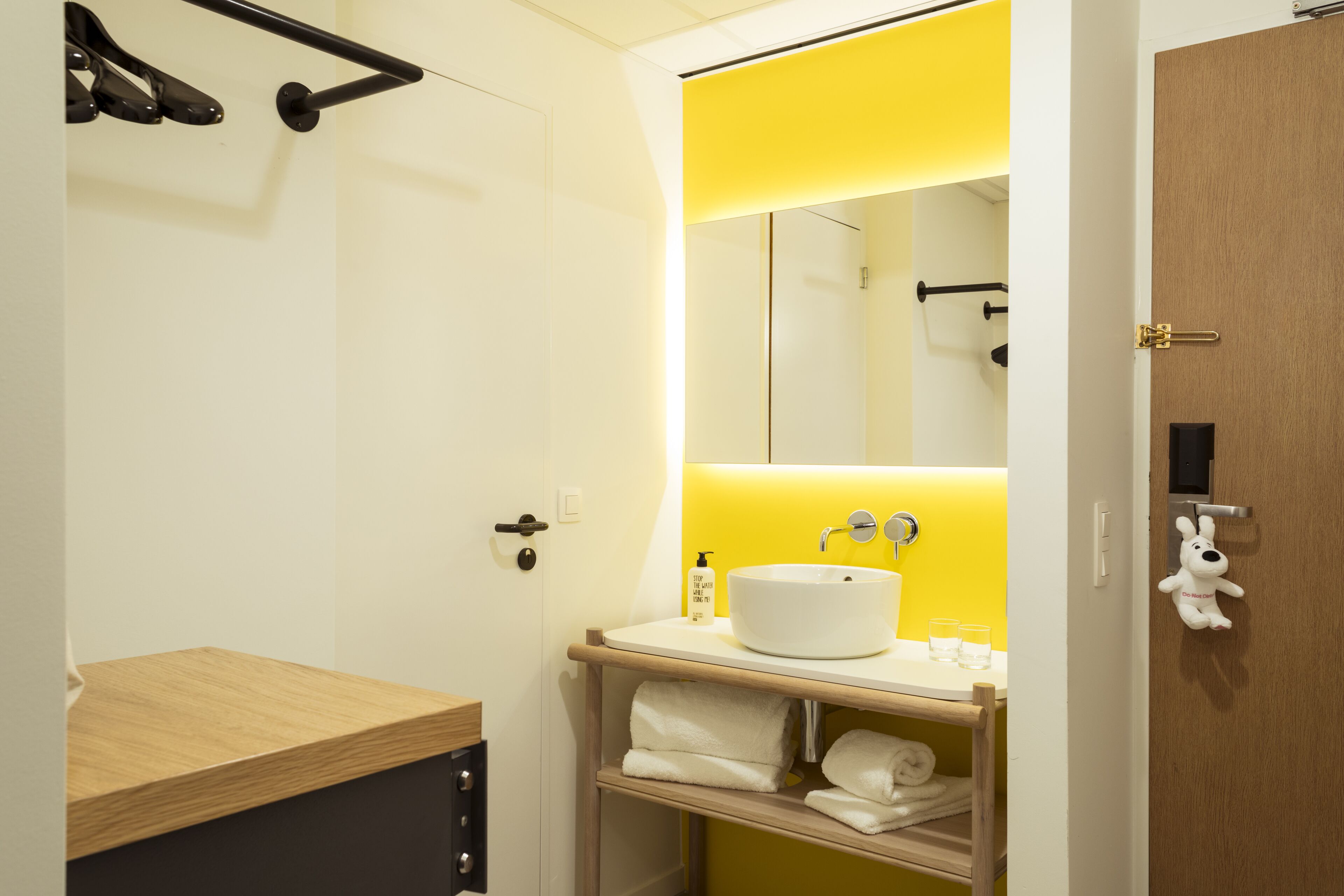 A modern bathroom with a wooden vanity, a white sink, and a yellow accent wall.