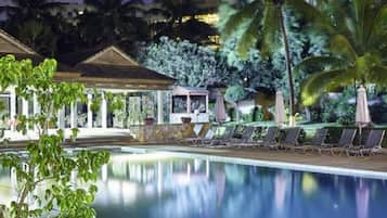 Un área de piscina nocturna con sillones reclinables y una zona de estar cubierta.