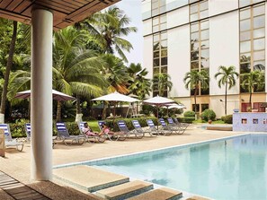 Un área de piscina con sillas de descanso y sombrillas, rodeada de palmeras y un edificio moderno.