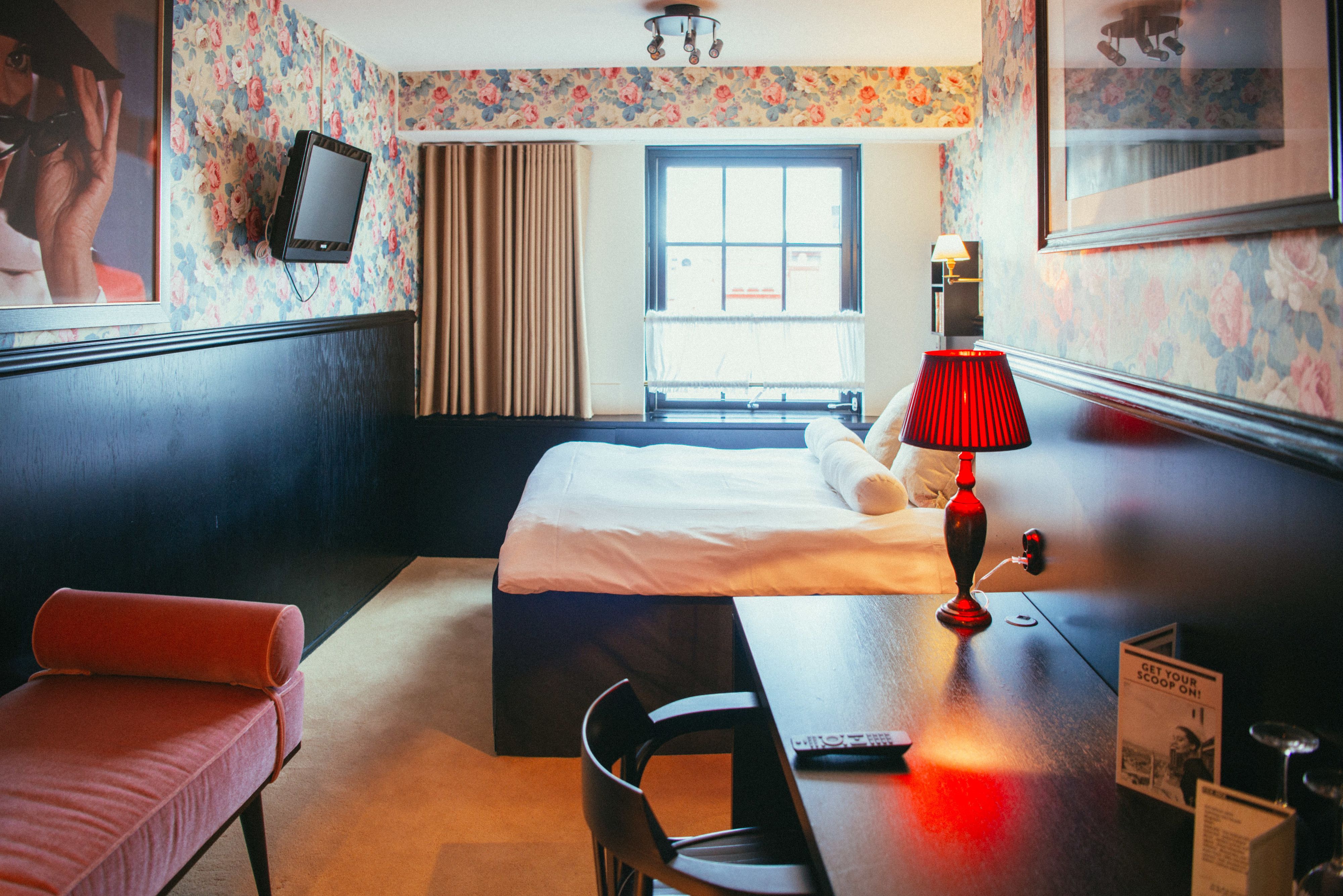 Una habitación de hotel con cama, escritorio con lámpara, silla, televisor en la pared y ventana con cortinas.
