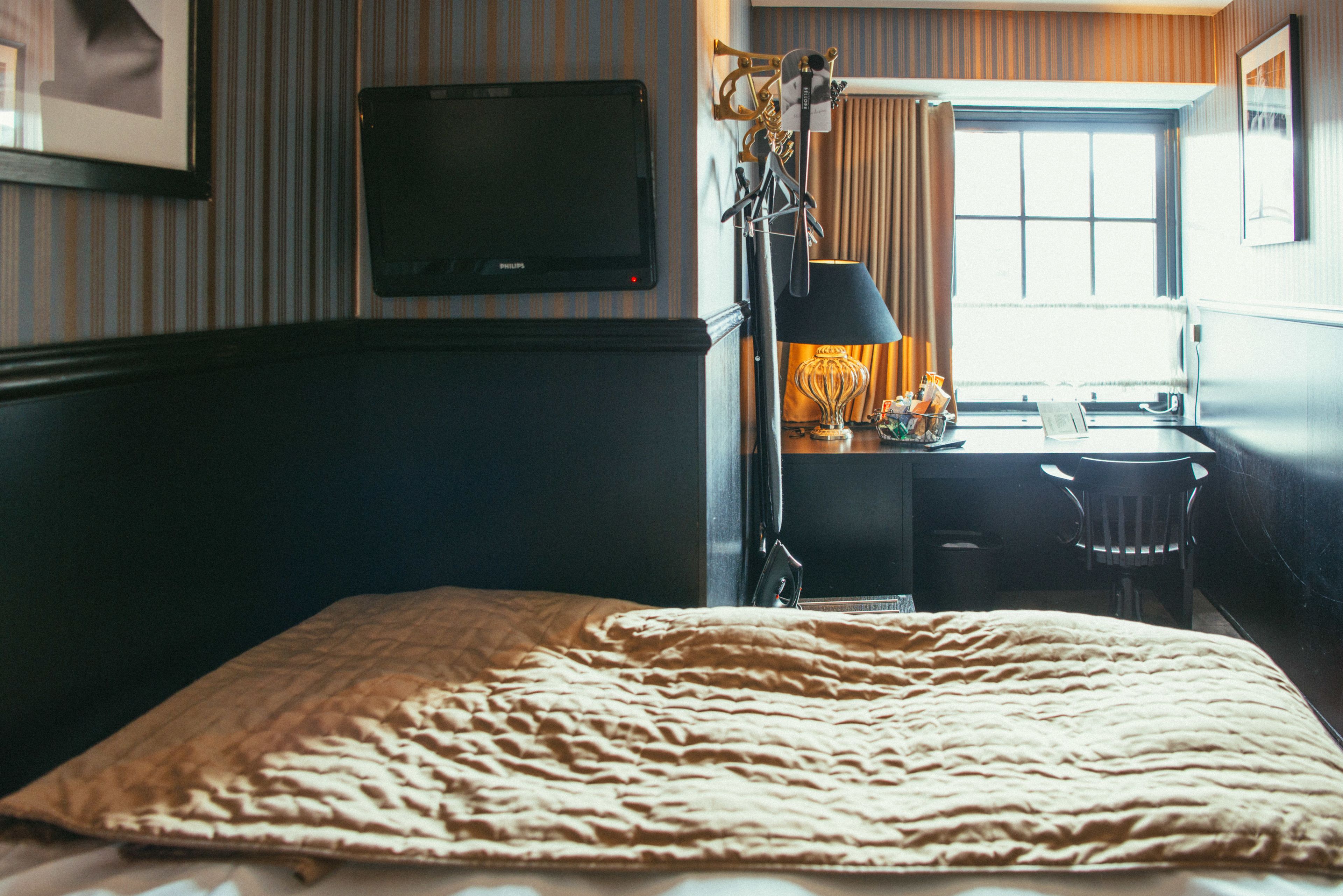 A hotel room with a bed, a desk with a lamp, a television, and a window with curtains.