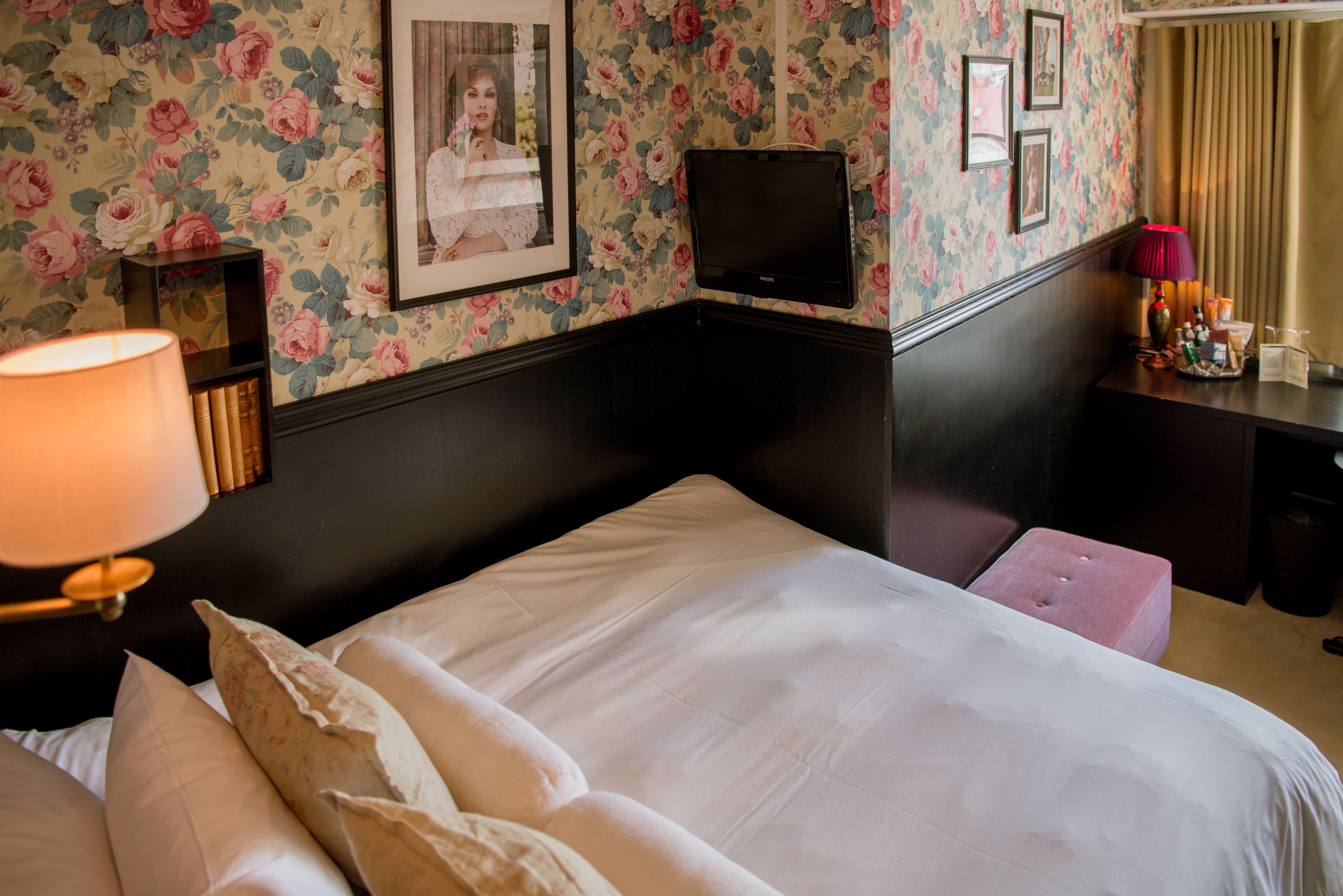 A hotel room with a bed, a bedside lamp, a flat-screen TV, a desk with a lamp, and a floral wallpaper.