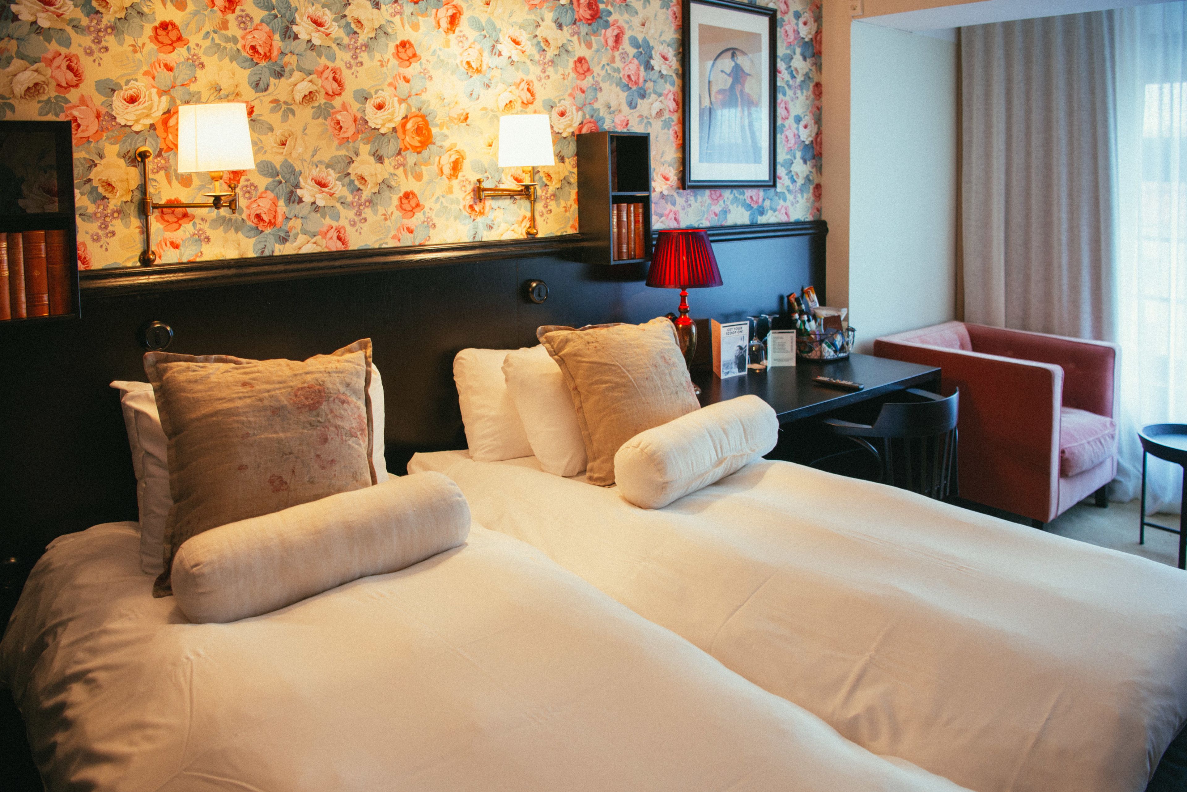 A hotel room with a large bed, two bedside lamps, a desk with a red lamp, a red chair, and a floral wallpaper.