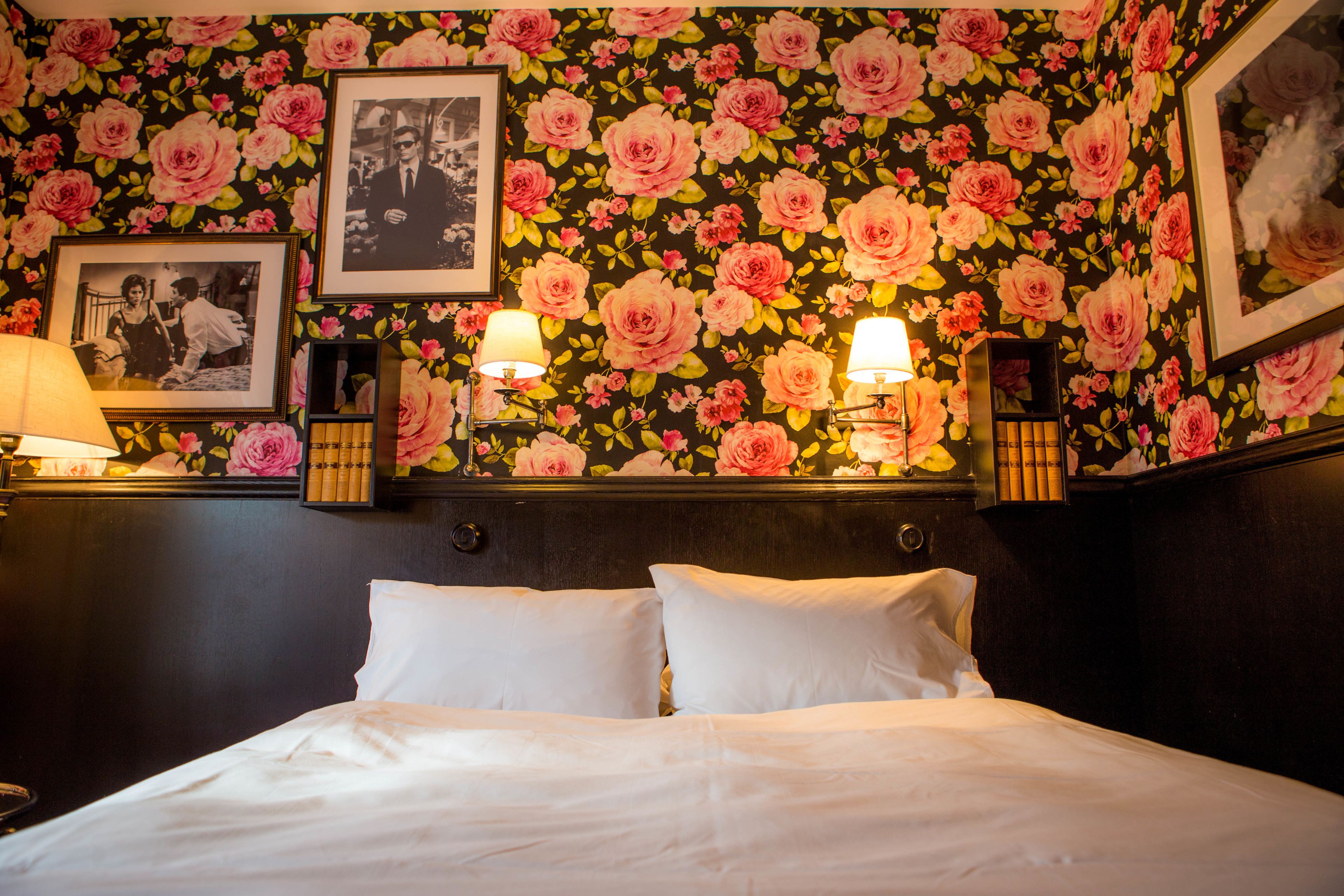Habitación de hotel con papel tapiz floral, una cama con ropa blanca, dos lámparas de noche y cuadros en la pared.