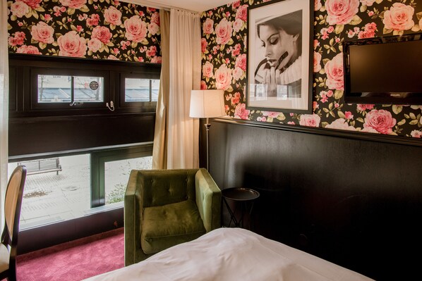 A hotel room with a bed, a green armchair, a small table, a window with a view of the street, and a television mounted on the wall.