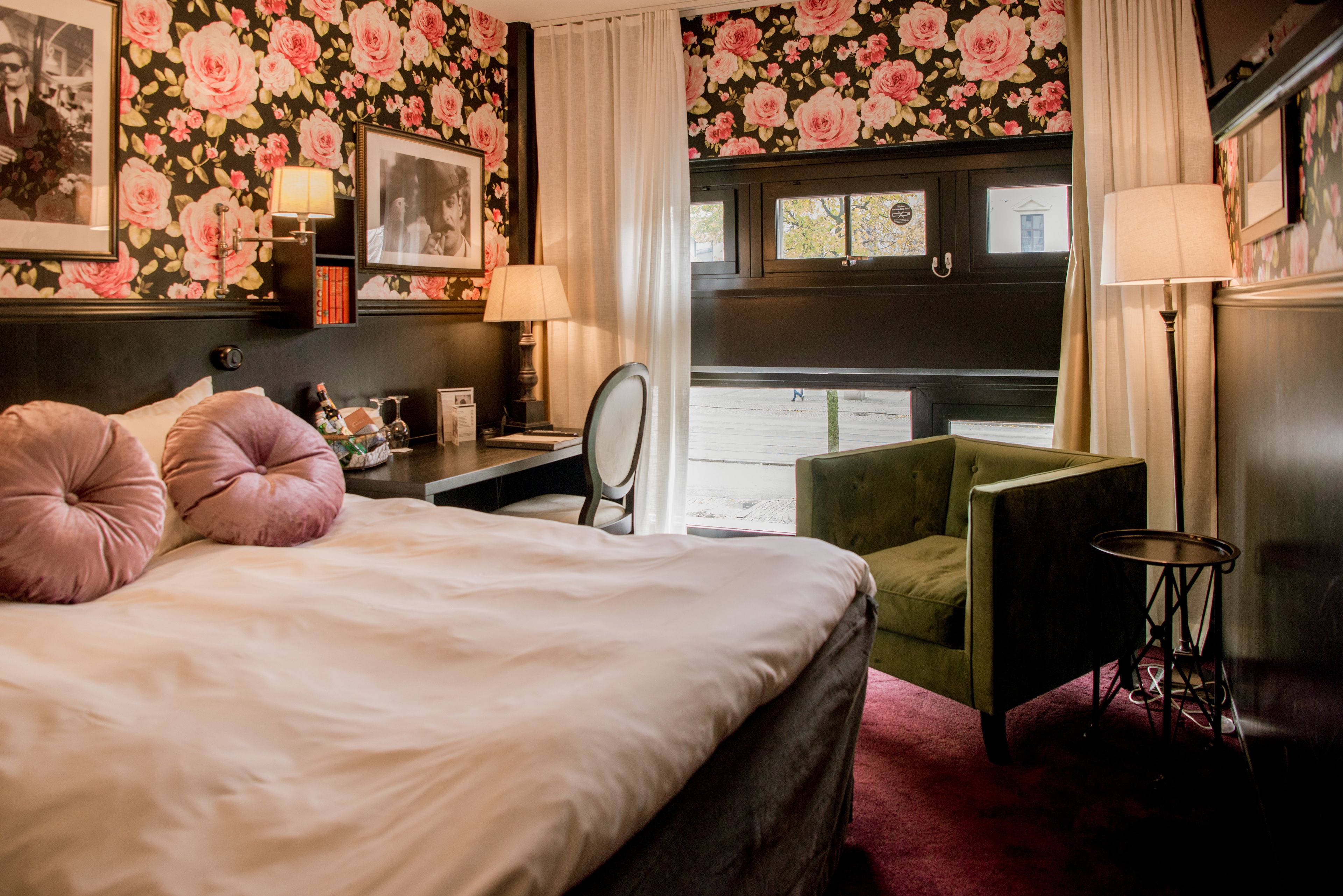 A hotel room with a bed, a green armchair, a desk with a lamp, and a window with curtains.