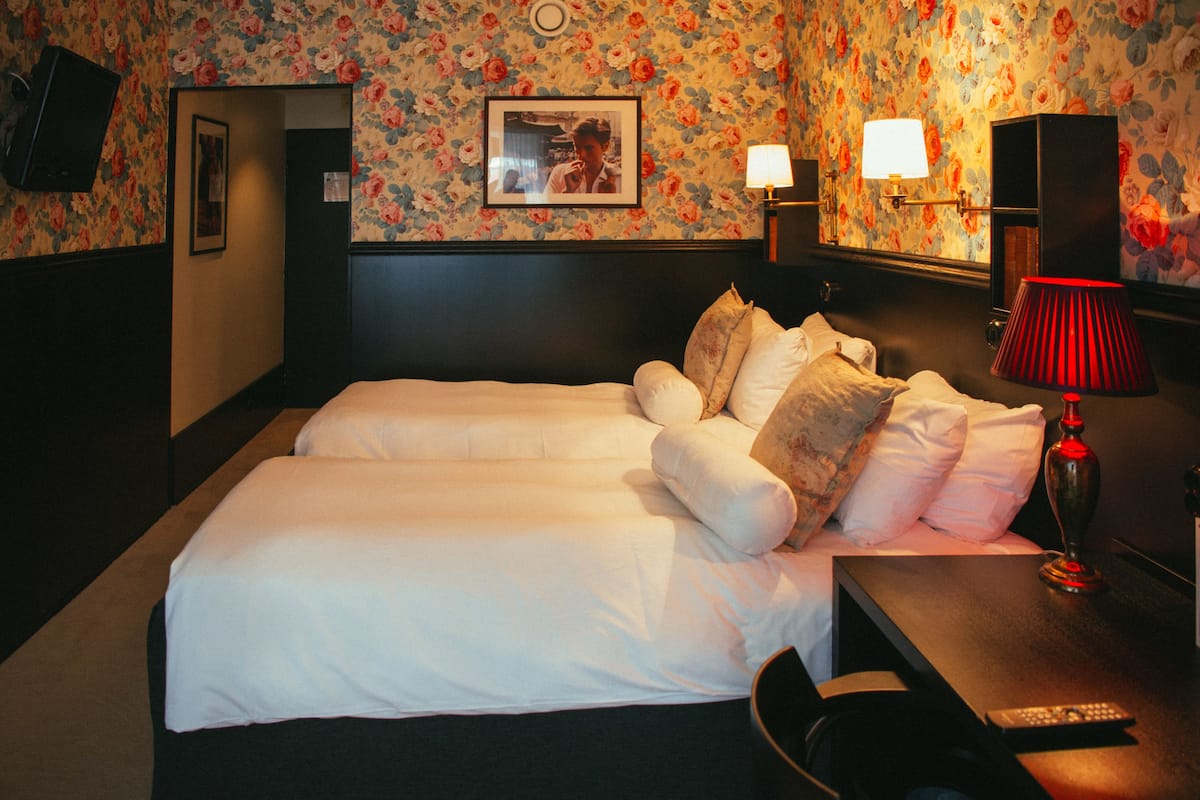 A hotel room with two beds, a desk, a chair, a TV, and a lamp.