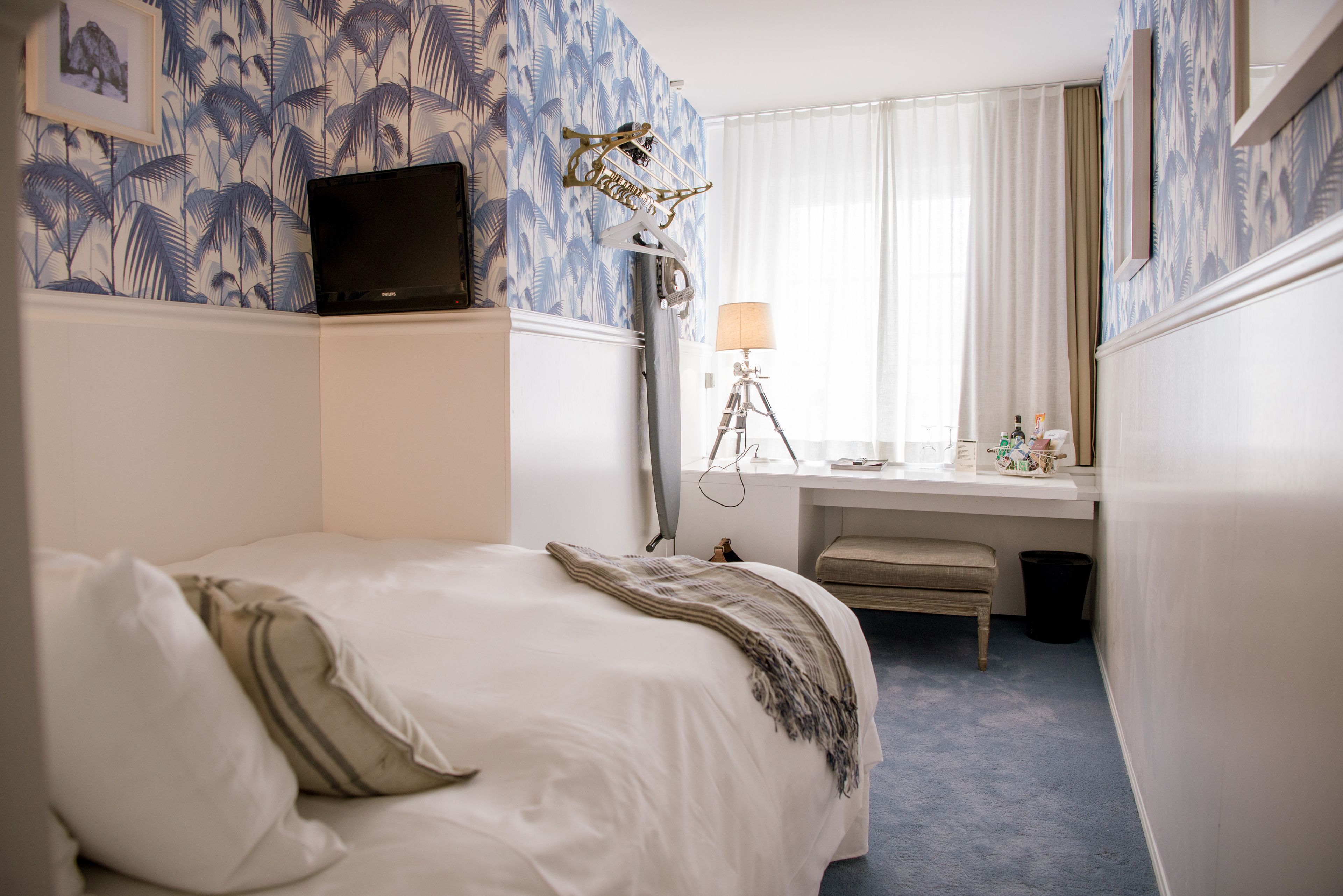 Ett hotellrum med en säng, ett skrivbord med en lampa, en bärbar dator som hänger på väggen och en papperskorg.