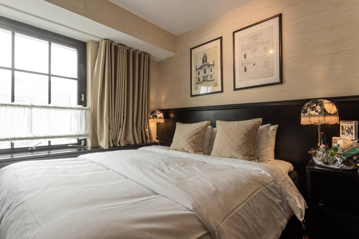 A neatly made bed with a white comforter and pillows, a window with curtains, and framed architectural drawings on the wall.