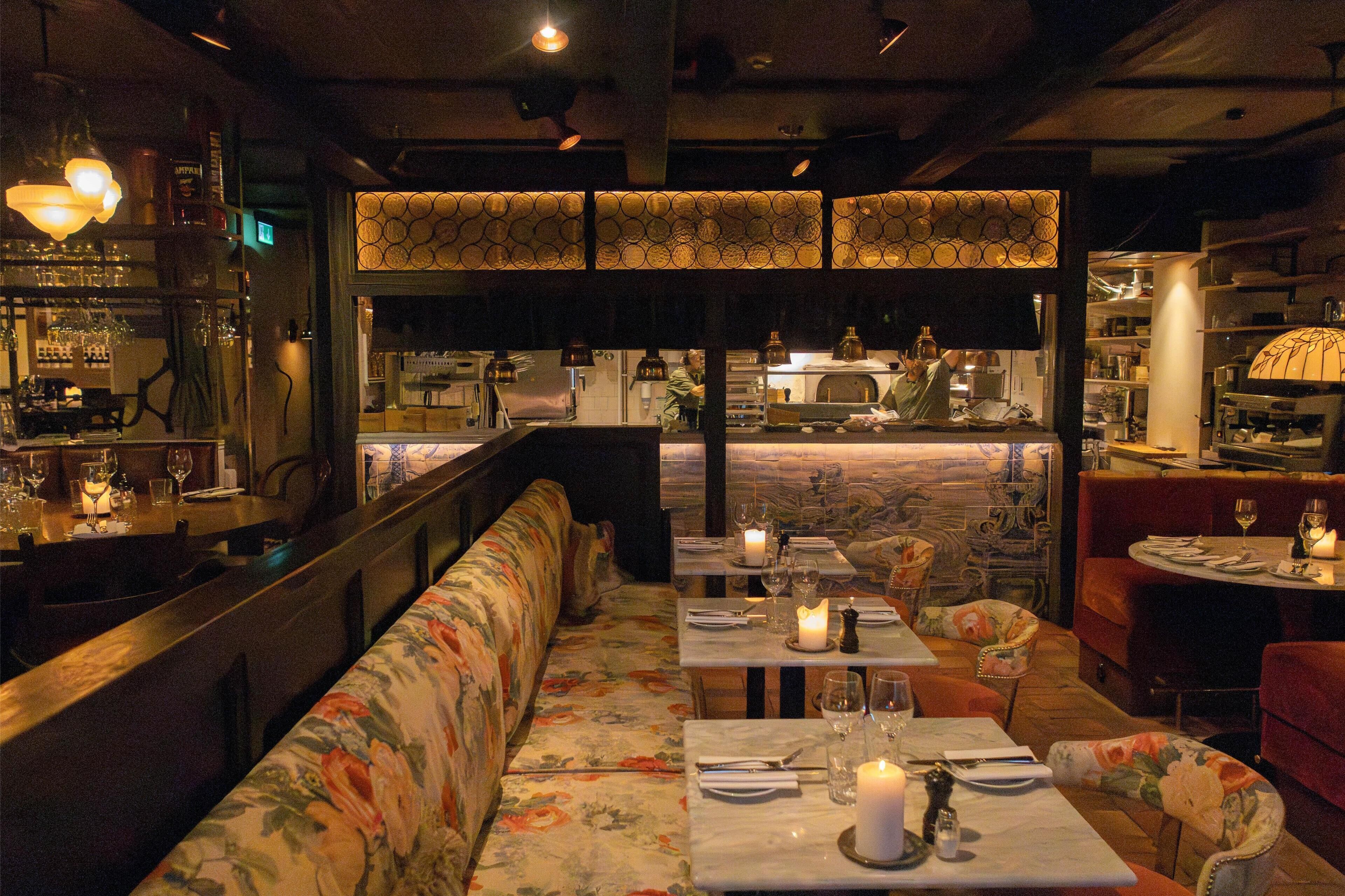 A restaurant interior with a long bench seating area, tables with candles, and a bar in the background.