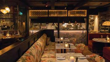 A restaurant interior with a long bench seating area, tables with candles, and a bar in the background.