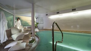 A modern indoor pool area with a seating area, a pool with a ladder, and a person in a robe. - Hotel Milton (Rimini)