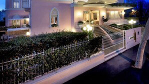 A pink building with a staircase leading to a balcony, surrounded by greenery and a fence. - Hotel Milton (Rimini)