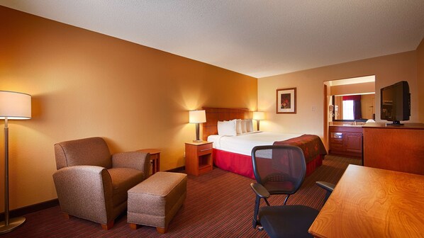 A hotel room with a bed, two armchairs, a desk, a chair, a TV, and a bathroom visible through an open door.