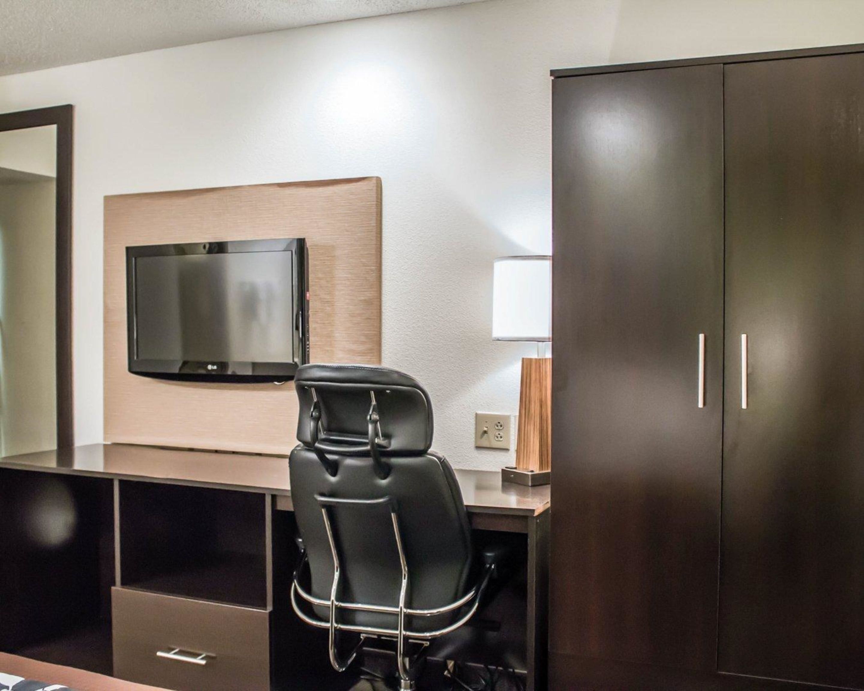 A hotel room with a flat-screen TV mounted on the wall, a desk with a chair, and a wardrobe.