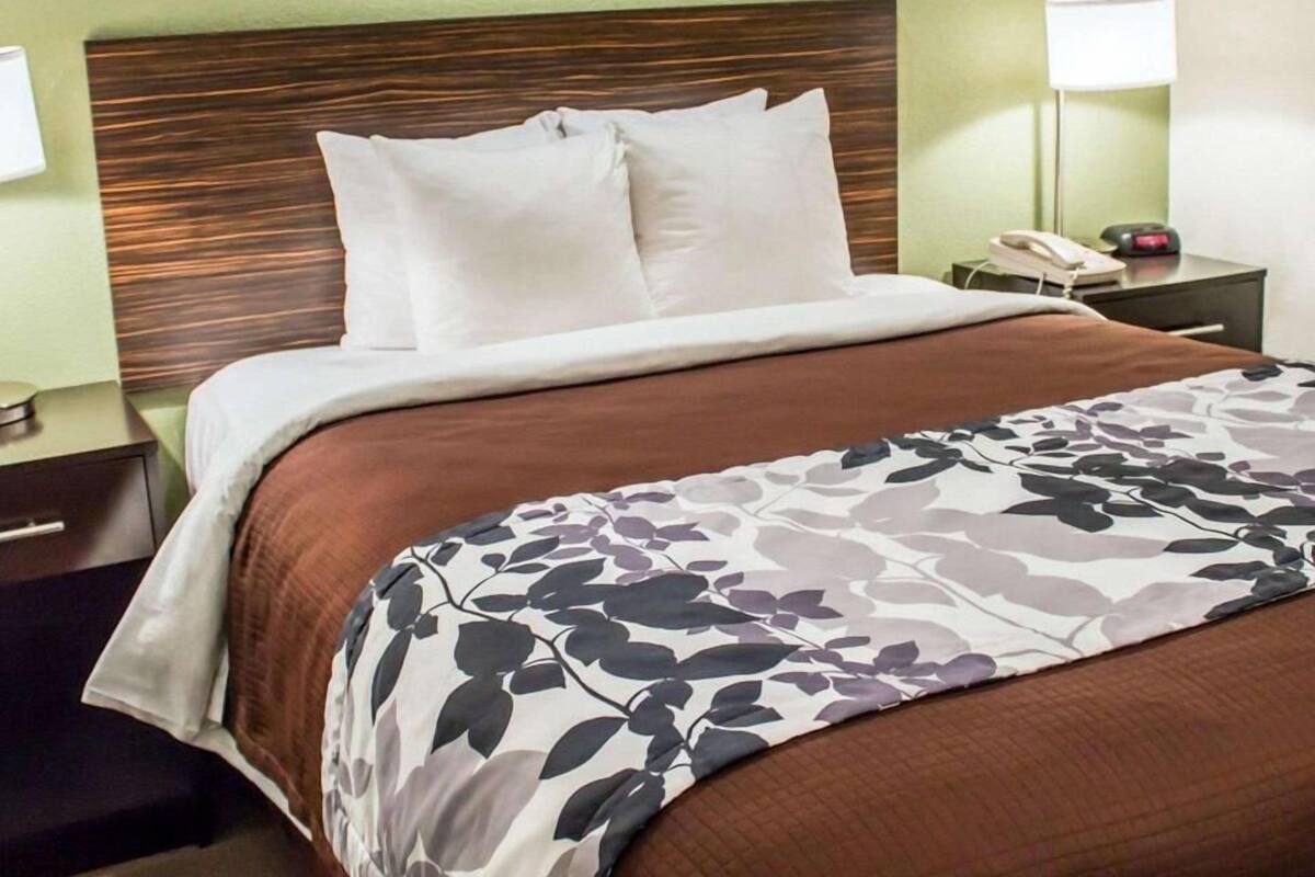 A neatly made bed with a brown and white floral bedspread, a wooden headboard, and a bedside table with a lamp and a phone.