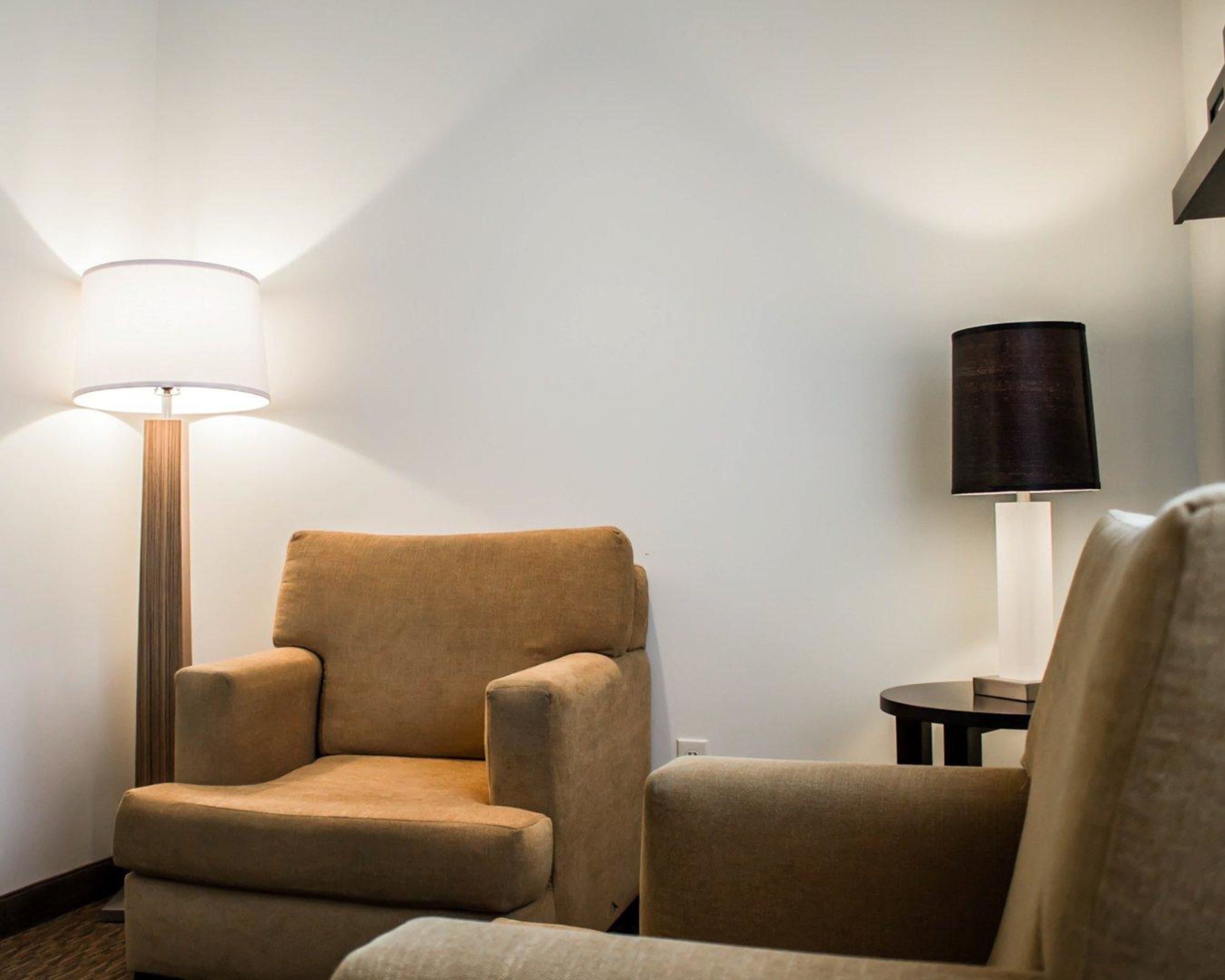Ett hotellrum med en fåtölj, en golvlampa och en bordslampa placerade framför en enkel vägg.