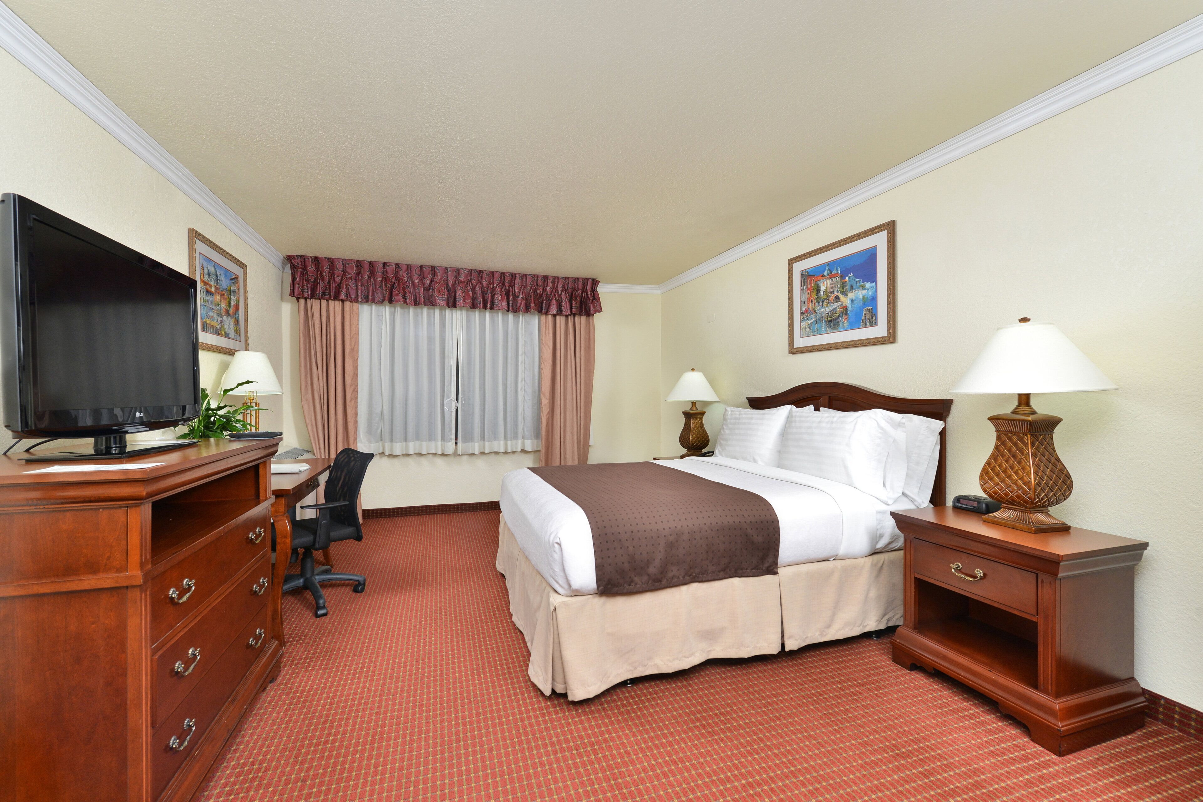 A hotel room with a bed, a desk with a television, a chair, two bedside tables with lamps, and a window with curtains.