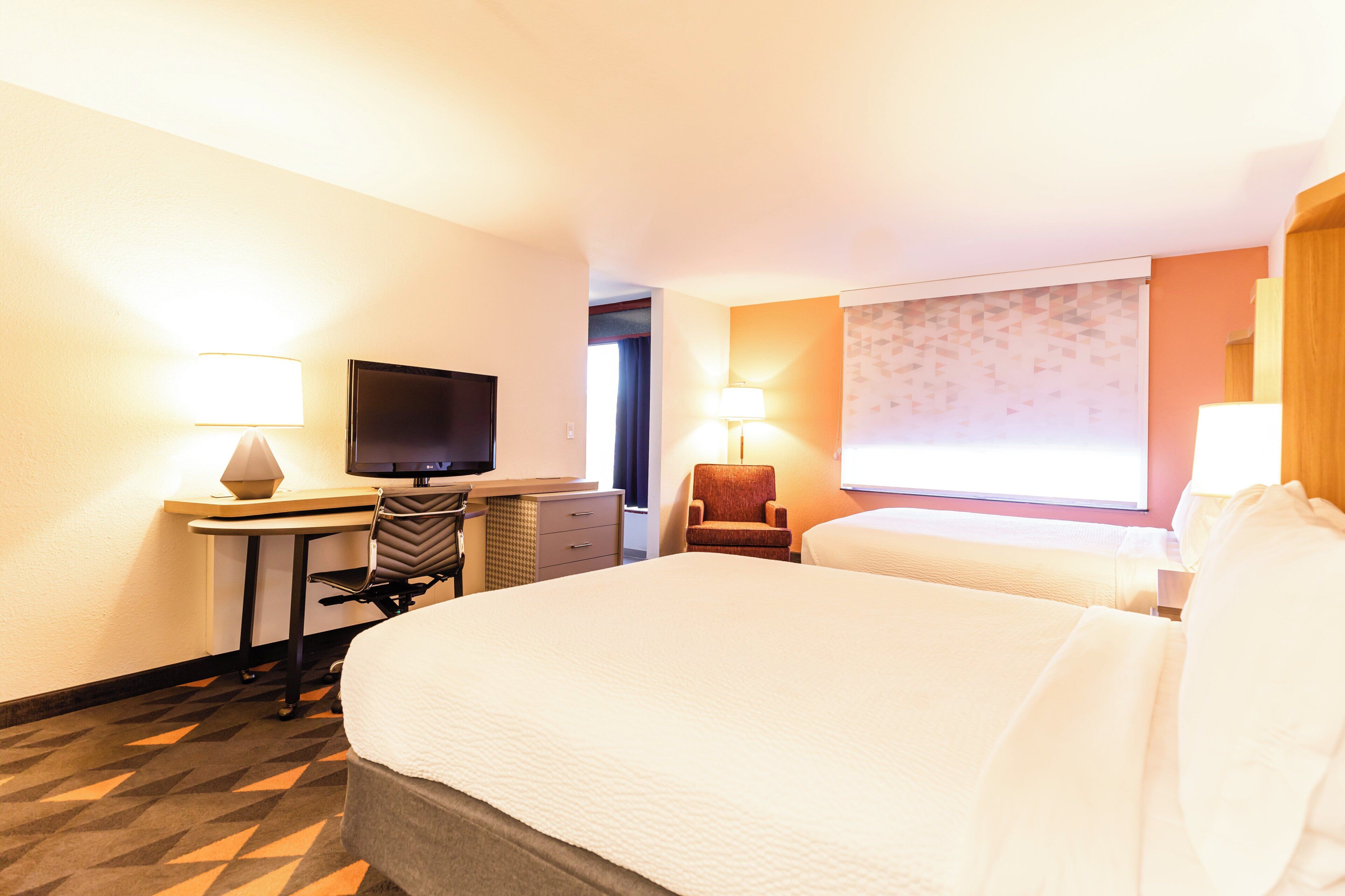 A hotel room with a large bed, a desk with a flat-screen TV, two lamps, and a window with a patterned curtain.