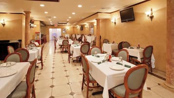 A hotel dining area with multiple tables and chairs, a tiled floor, and a television mounted on the wall.