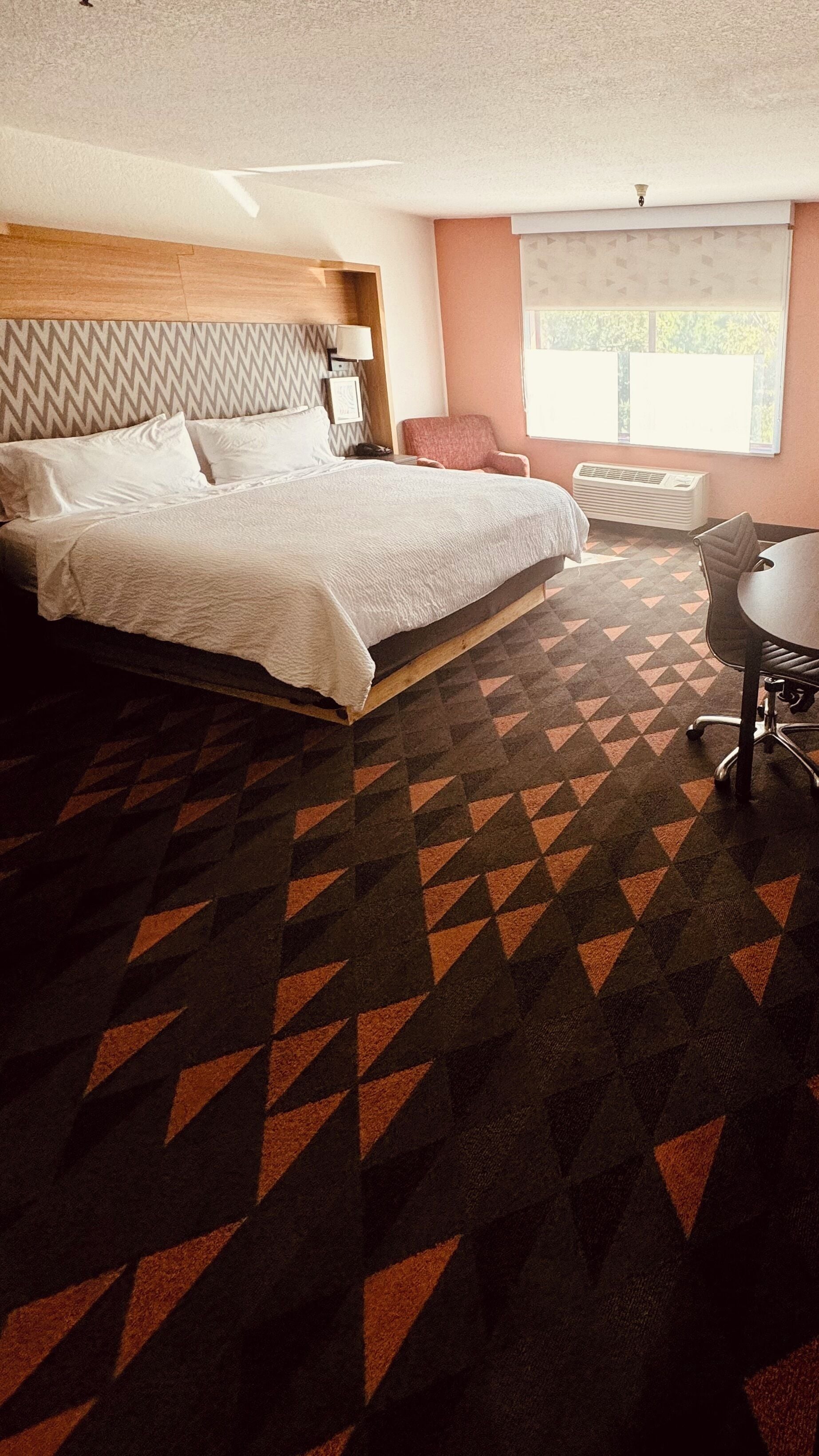 A hotel room with a large bed, a desk, and a chair. There is a window with blinds and a ceiling light.