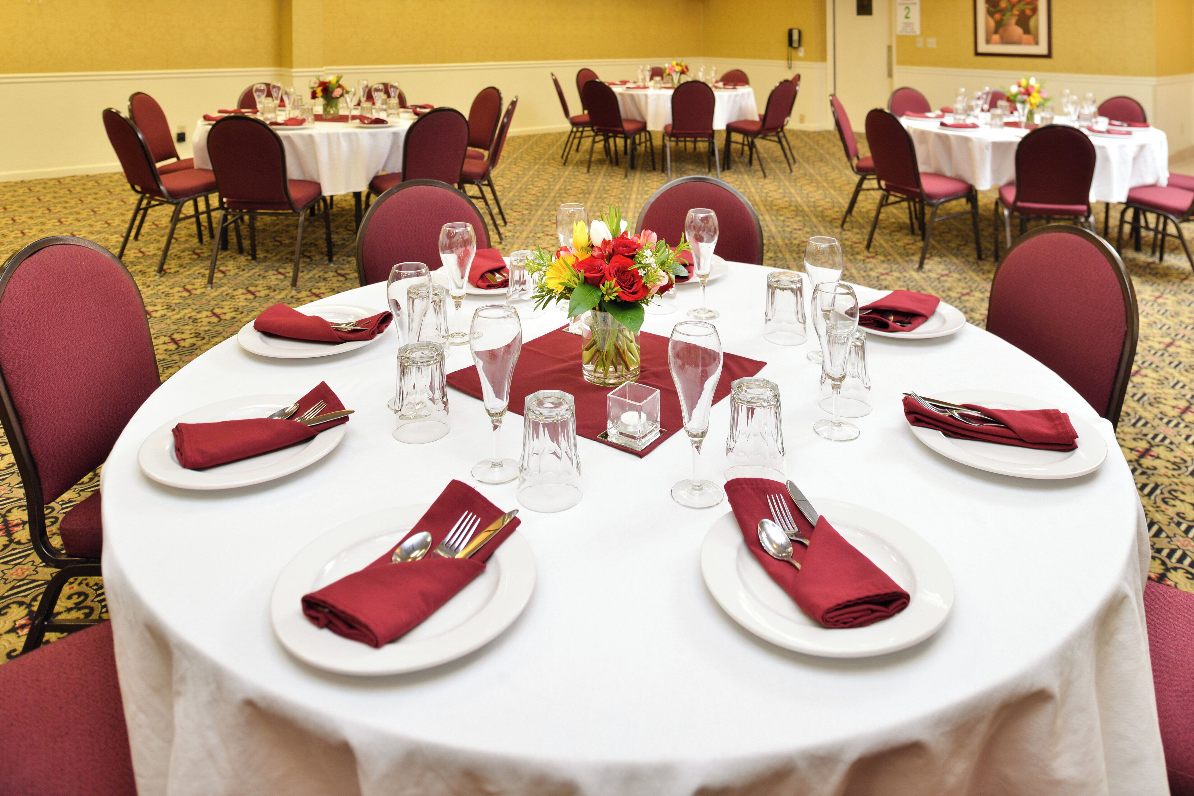 A banquet hall with round tables set for dining, each with white tablecloths, red napkins, and clear glassware.