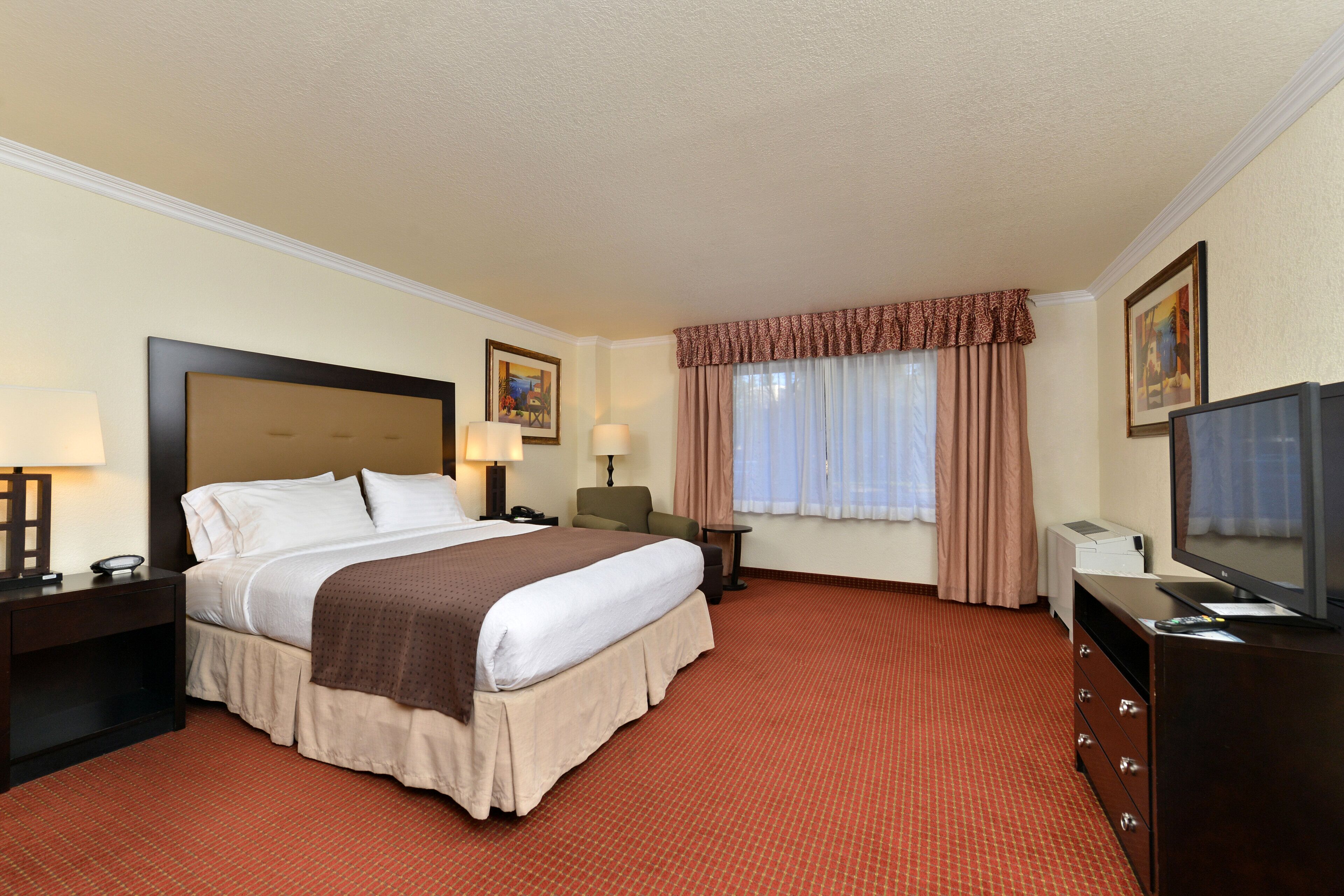 A hotel room with a large bed, bedside lamps, a desk with a lamp, a television, and a window with curtains.