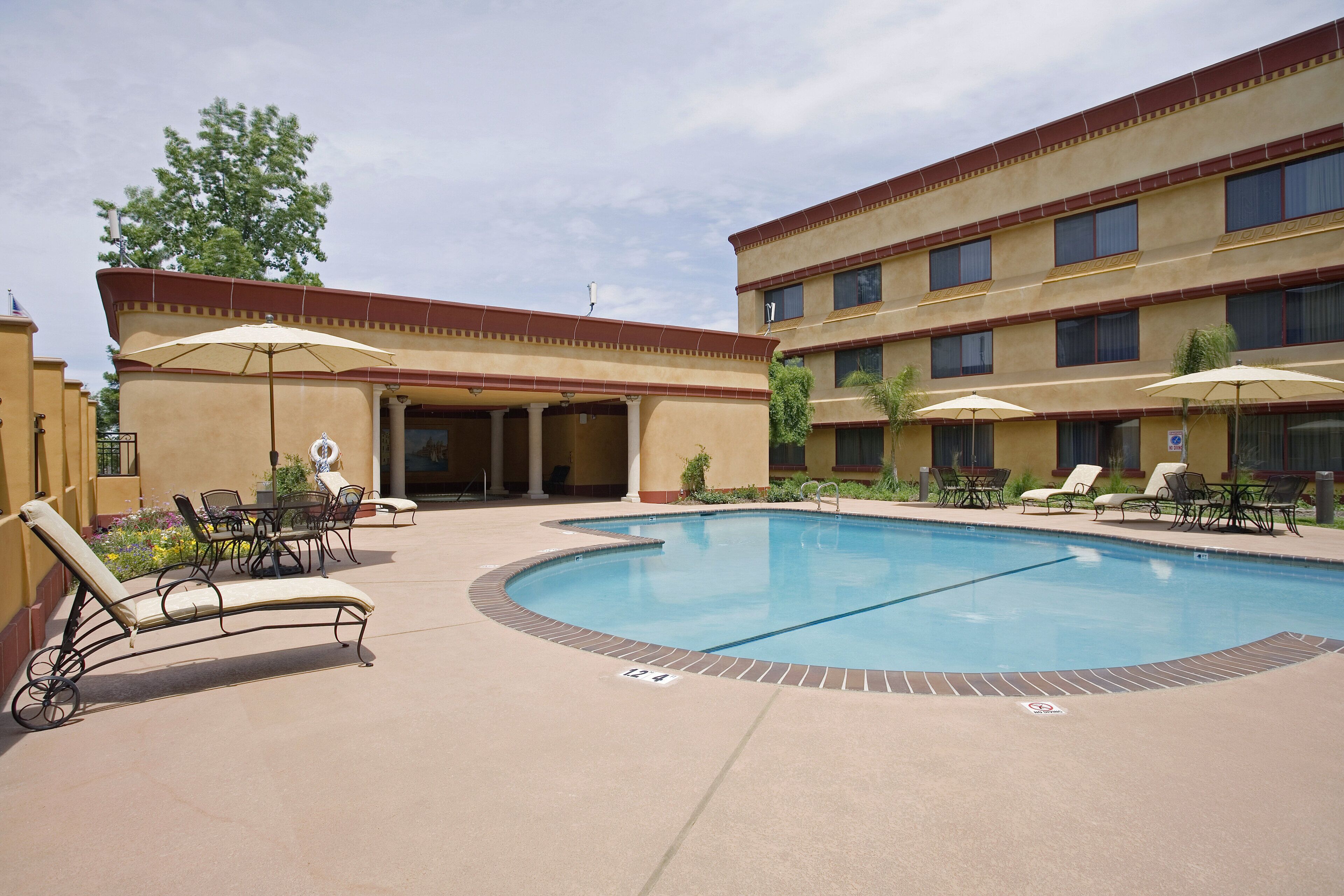 A hotel pool area with lounge chairs and umbrellas, a covered seating area, and a building with multiple windows.