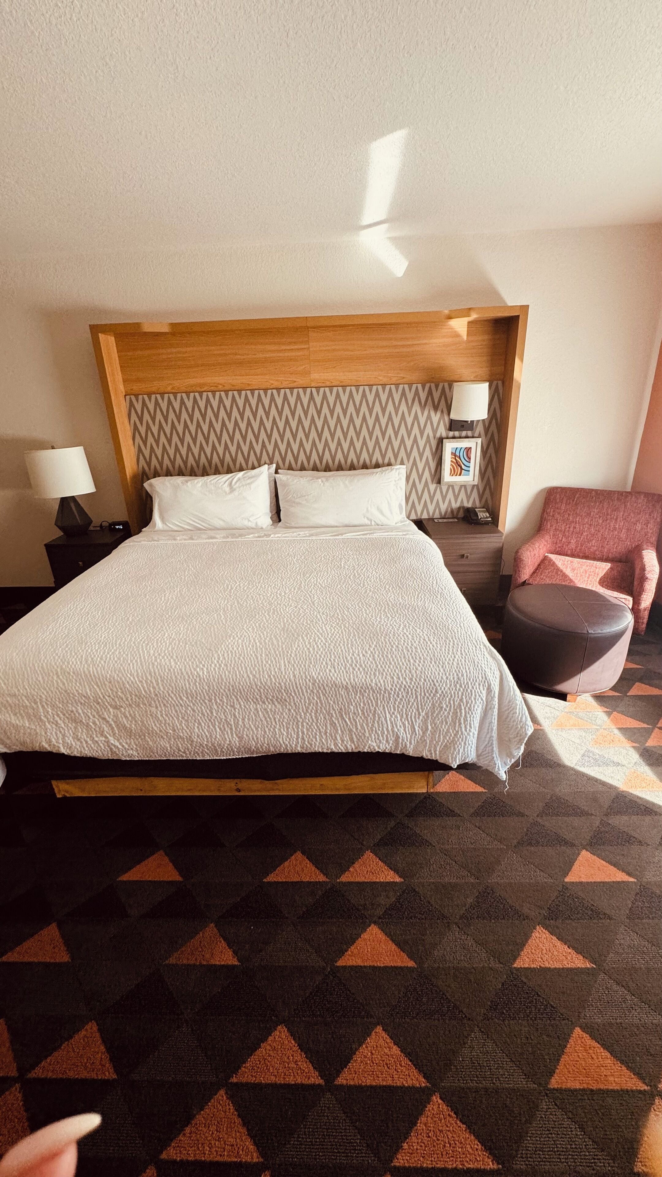 Habitación de hotel con una cama grande, cabecera de madera, una silla, una lámpara y una alfombra estampada.